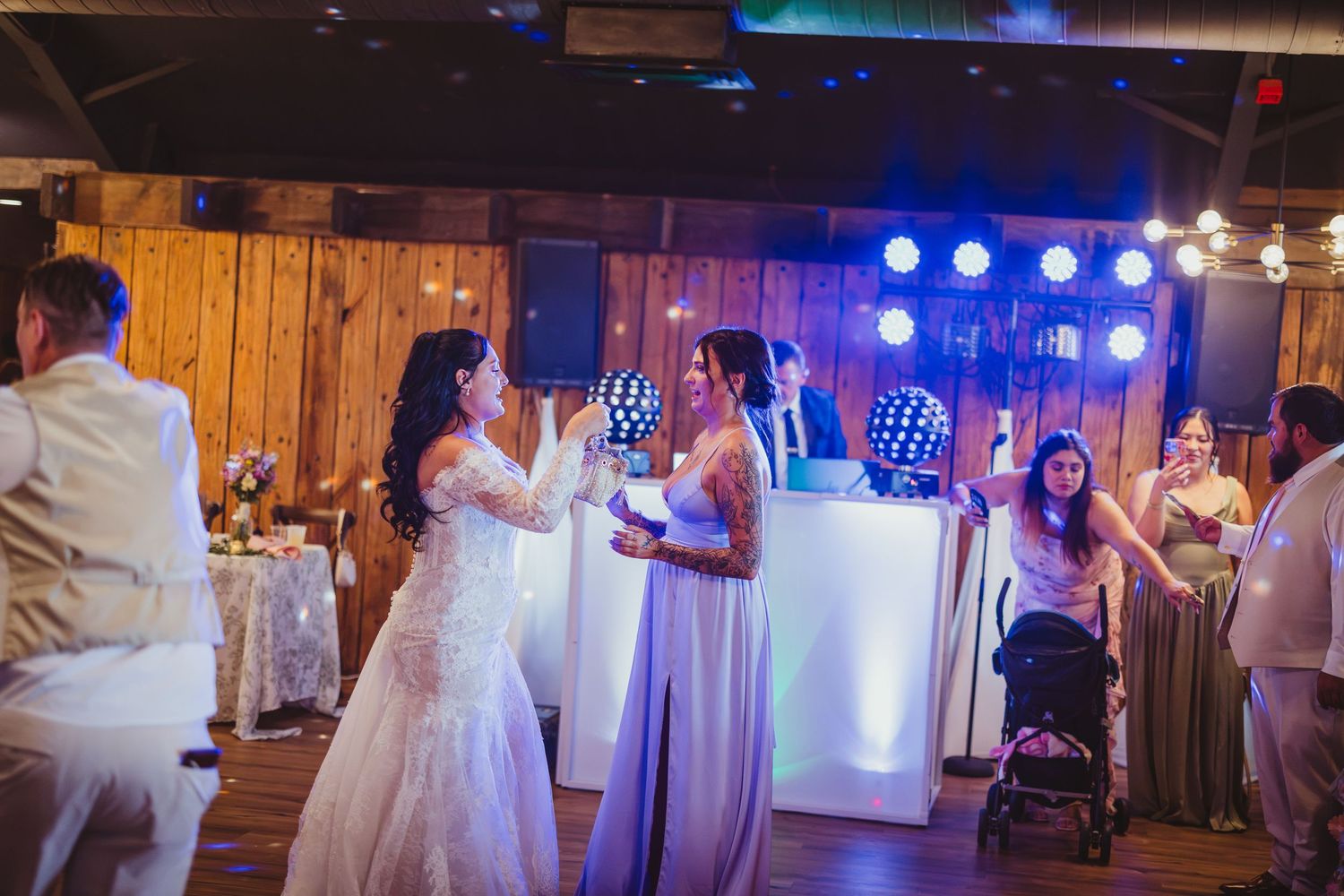 Bride and bridesmaid dancing joyfully at a rustic barn wedding reception with blue stage lights.
