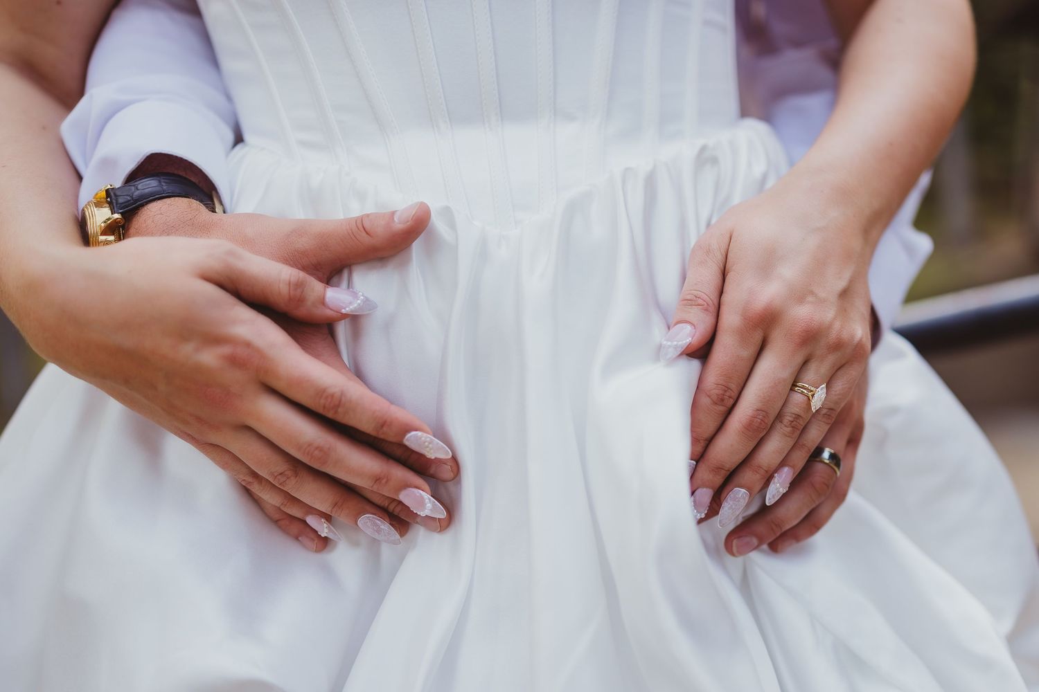 Close-up of two people's hands resting on a white wedding dress, wearing gold rings and a watch.