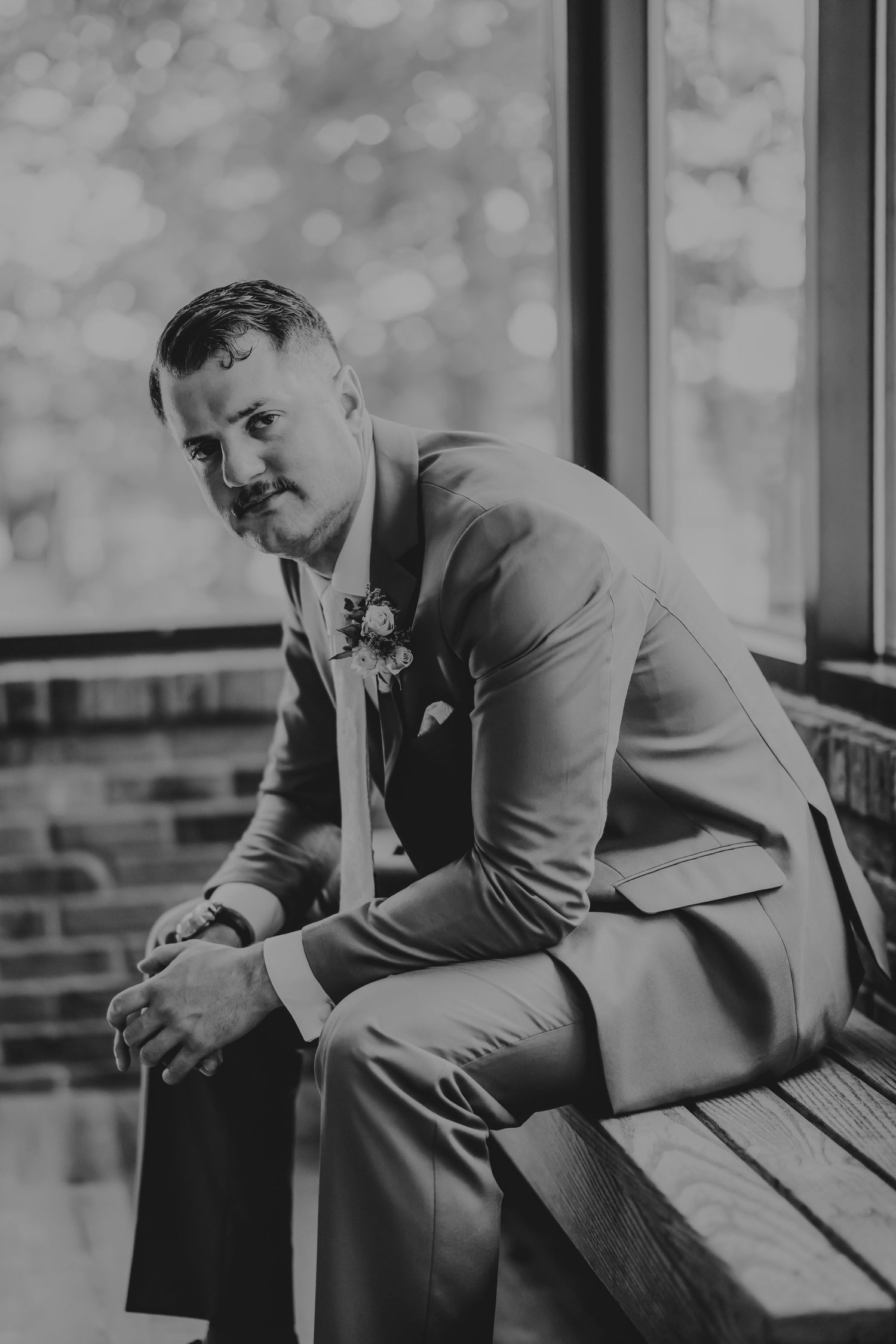 Black and white portrait of a groom in a suit with boutonniere sitting on a bench near a window.