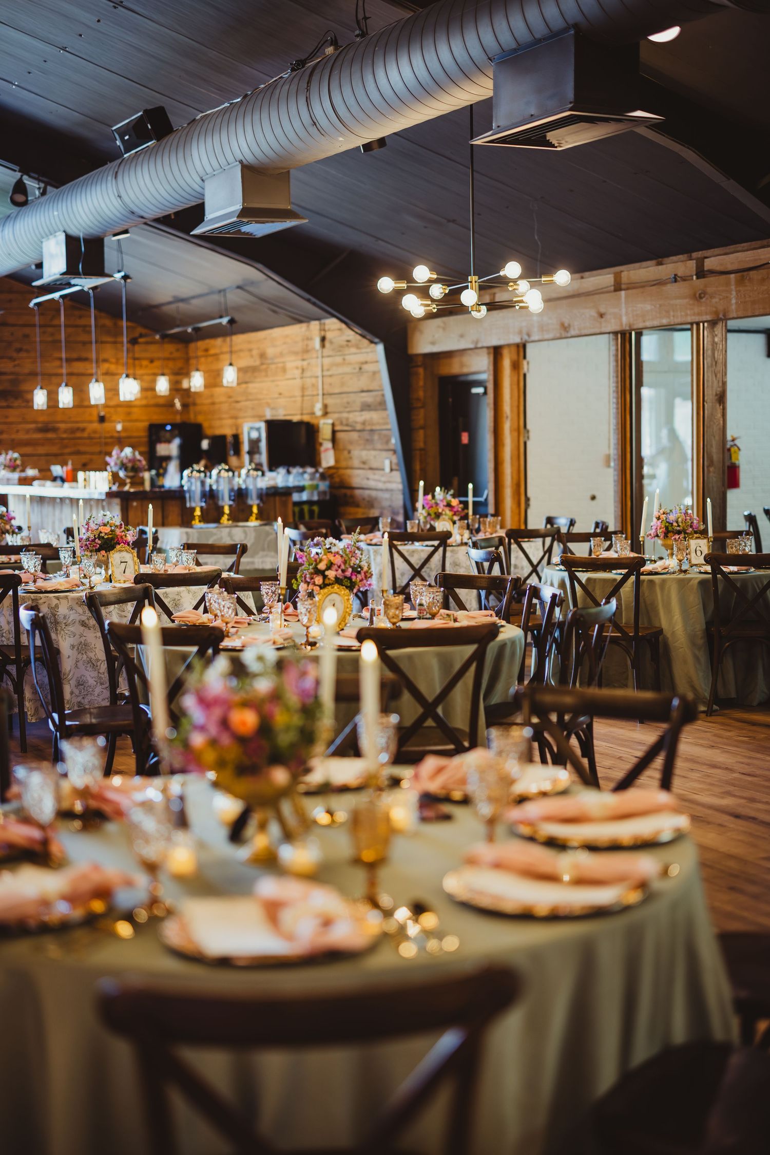 Elegant rustic wedding reception hall with round tables, floral centerpieces, candles, and warm Edison bulb lighting.