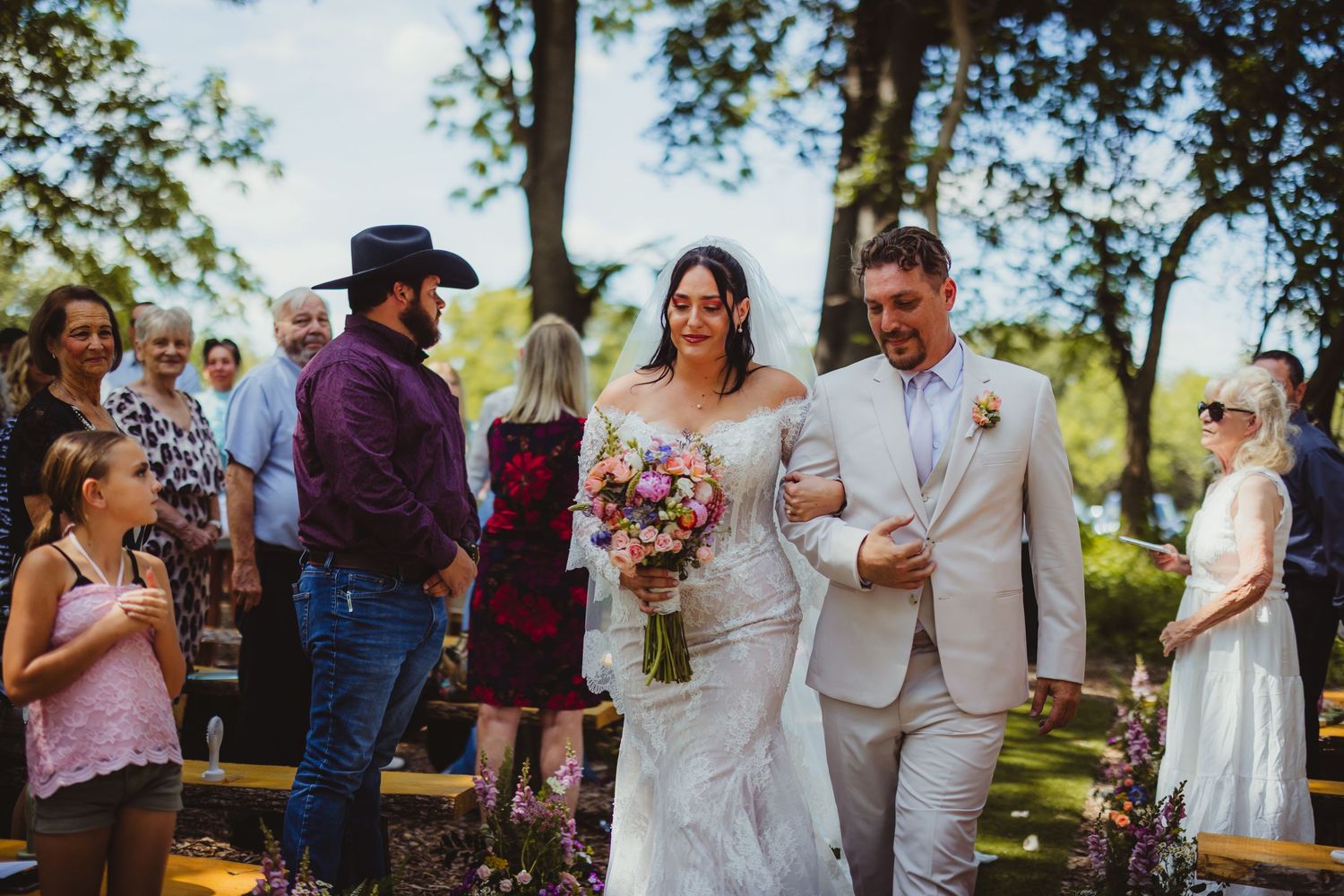 Bride and groom walk down the aisle at an outdoor wedding ceremony, bride holding a colorful bouquet.
