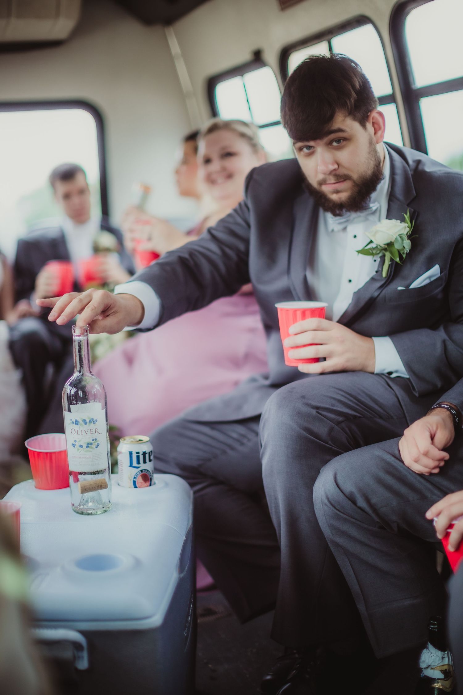 Groomsman in gray suit handles beverages while seated on wedding transportation.