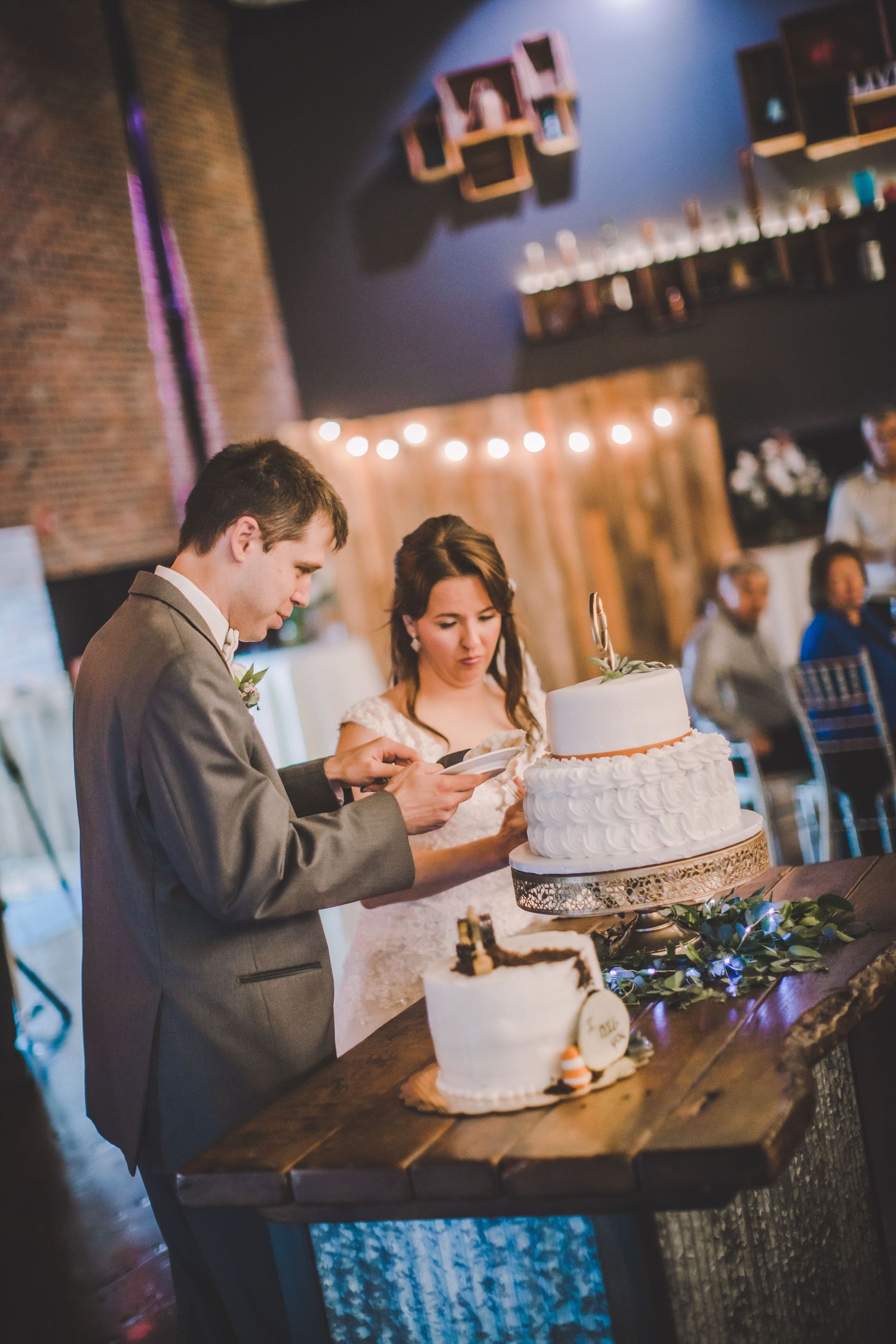 Wedding couple enjoys cake cutting tradition at reception with string lights and industrial decor.