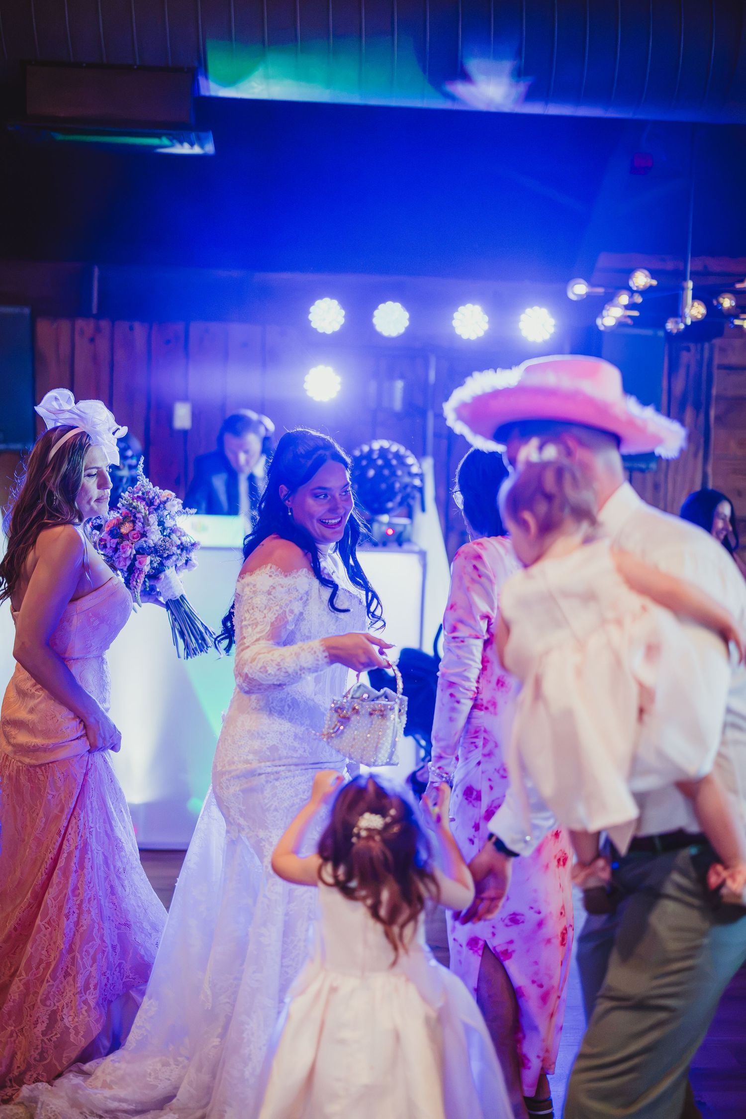 Bride holding bouquet dancing with guests including flower girl and man in pink cowboy hat at wedding reception.