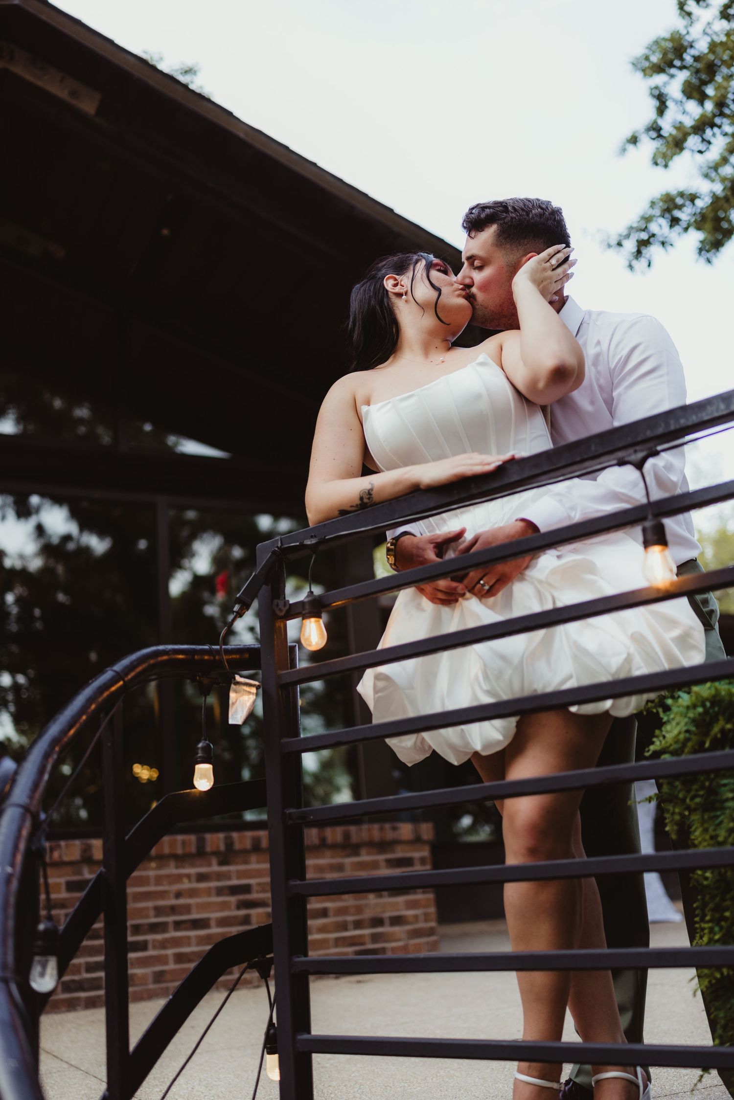 Bride and groom sharing a romantic kiss on a balcony with string lights, bride wearing a short white strapless dress.