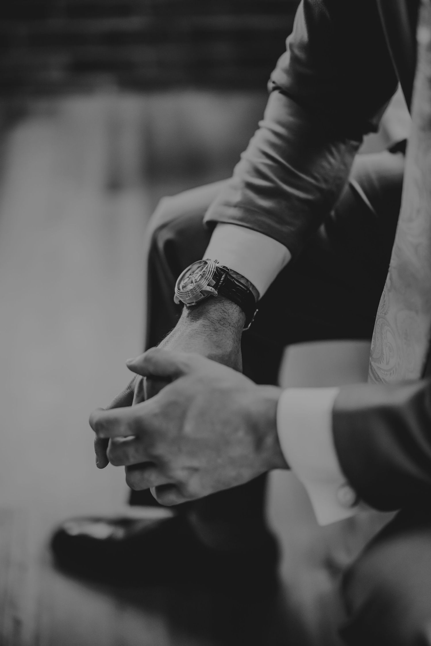 Black and white close-up of a man in a suit with a luxury wristwatch, hands clasped elegantly.