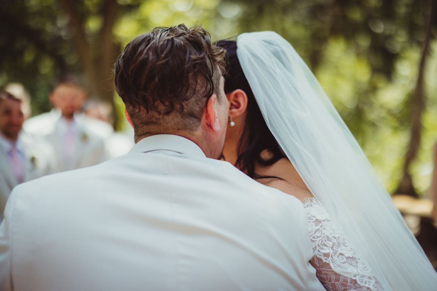 Bride and groom sharing first kiss at outdoor wedding ceremony, bride wearing white veil and lace dress.