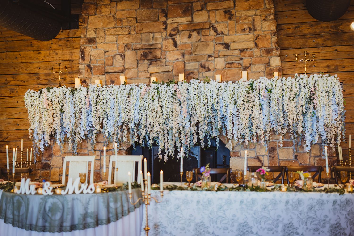 Elegant wedding sweetheart table with cascading white floral backdrop, candles, and Mr. & Mrs. sign in rustic venue.