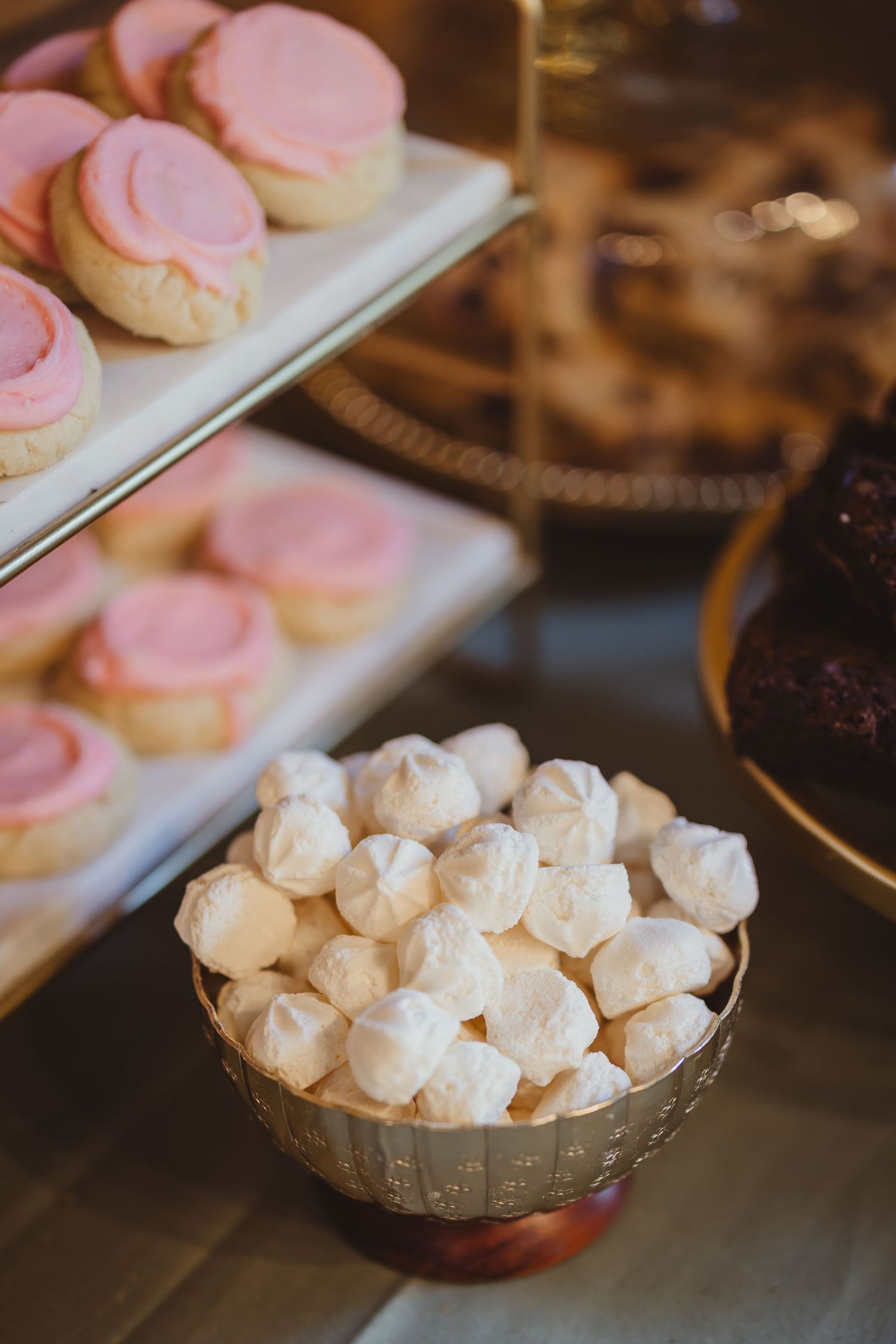 Gold bowl of white meringue cookies beside pink frosted sugar cookies on a tiered dessert display.
