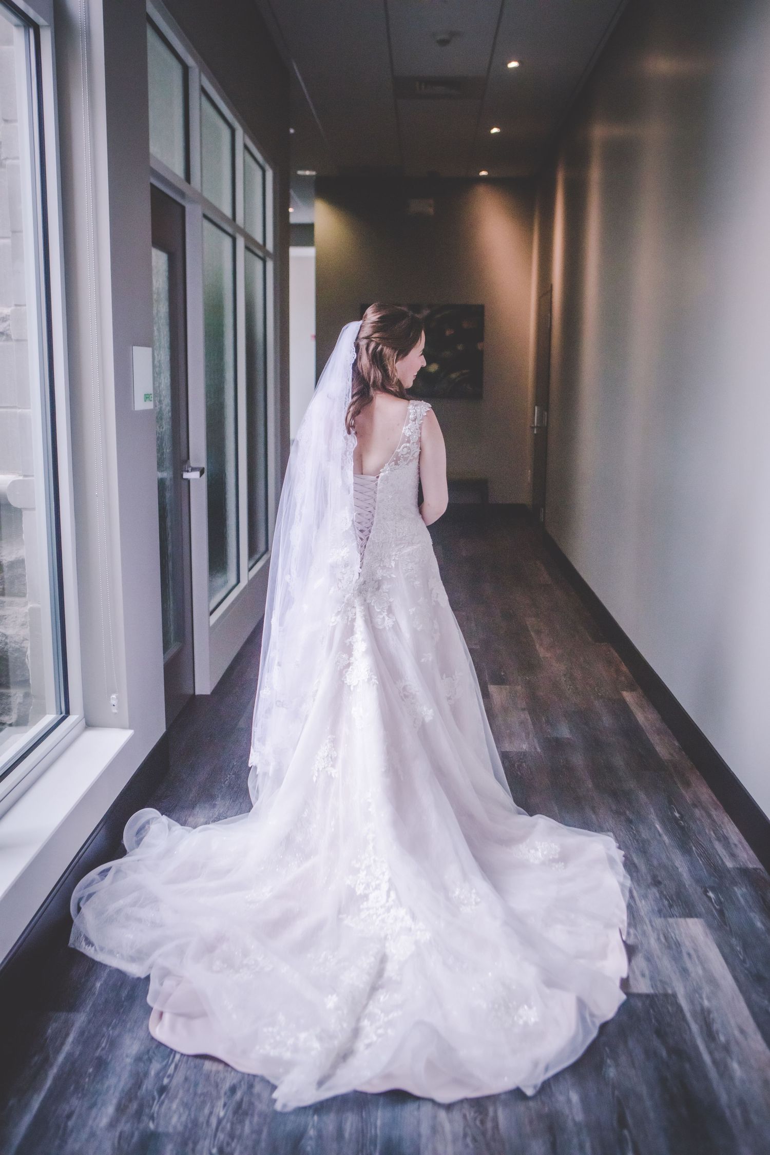 A flowing white lace wedding gown with long train photographed from behind in a modern hallway.