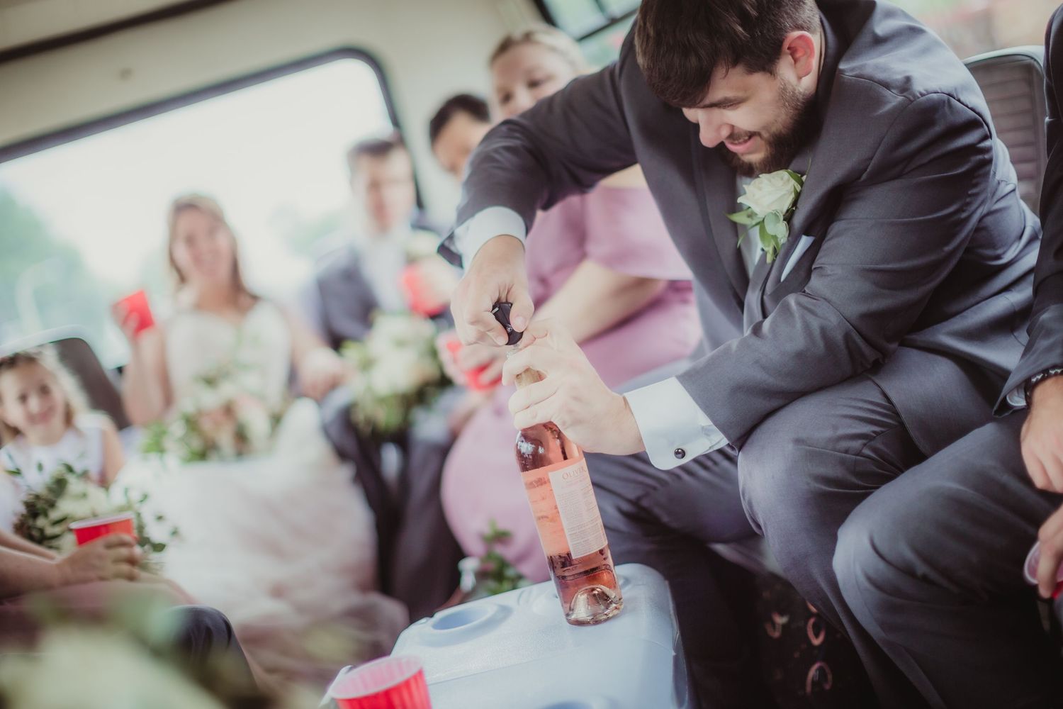 Wedding party member in suit carefully pours beverages during bus transportation.