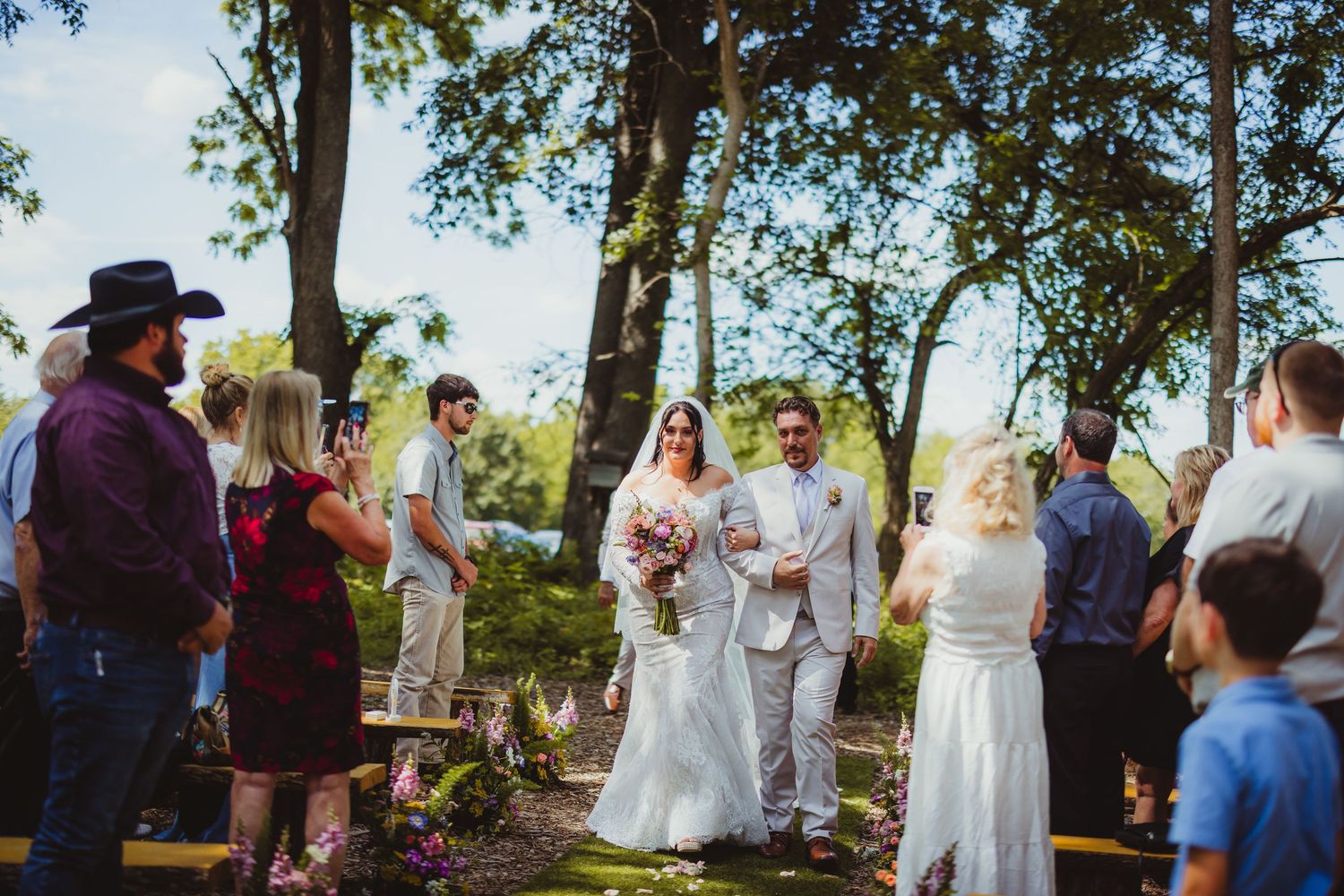 Bride and groom walk down the aisle at an outdoor wedding ceremony surrounded by guests and tall trees.