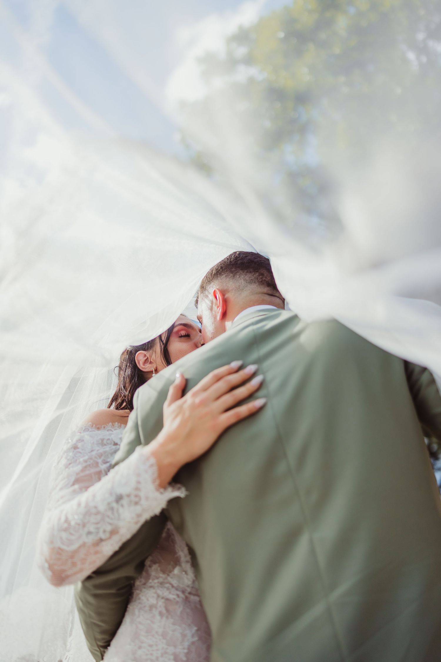 Bride and groom sharing a romantic embrace under a flowing white veil outdoors on their wedding day.