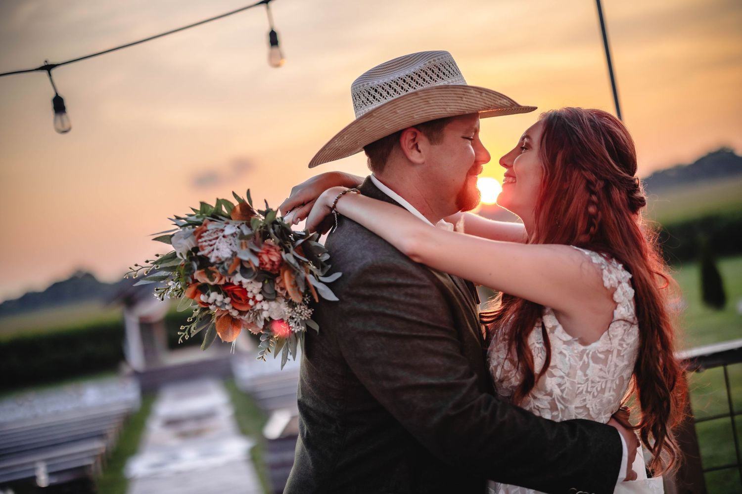 A couple sharing a romantic kiss at sunset amid a rustic farm setting with string lights overhead and floral decorations.