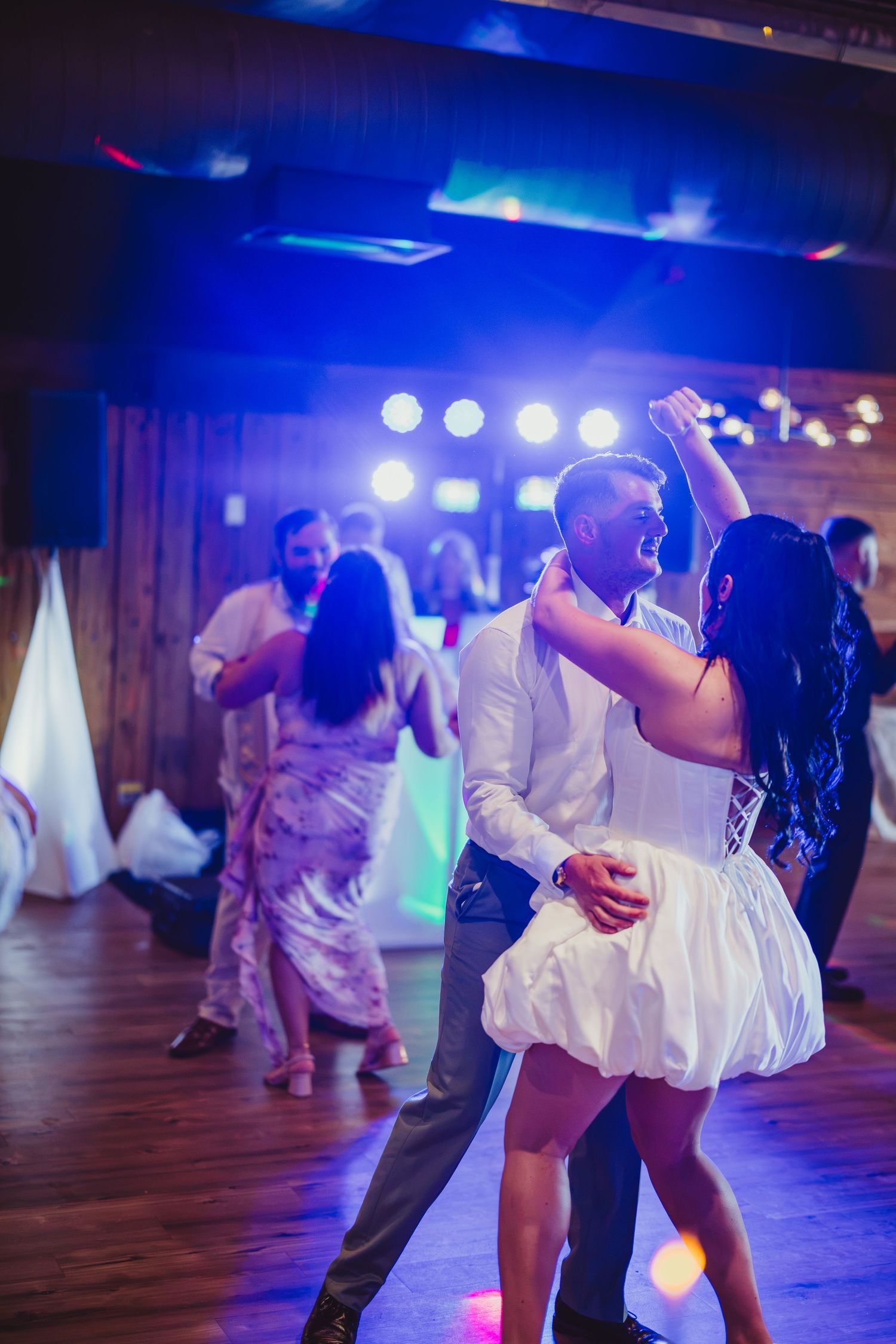 Couple dancing closely at a wedding reception with colorful stage lights and guests in the background.