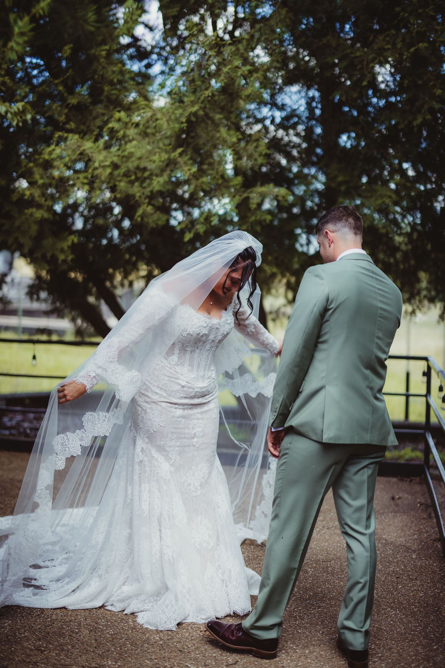 Bride in lace gown and veil shares a joyful moment with groom in sage suit outdoors on their wedding day.