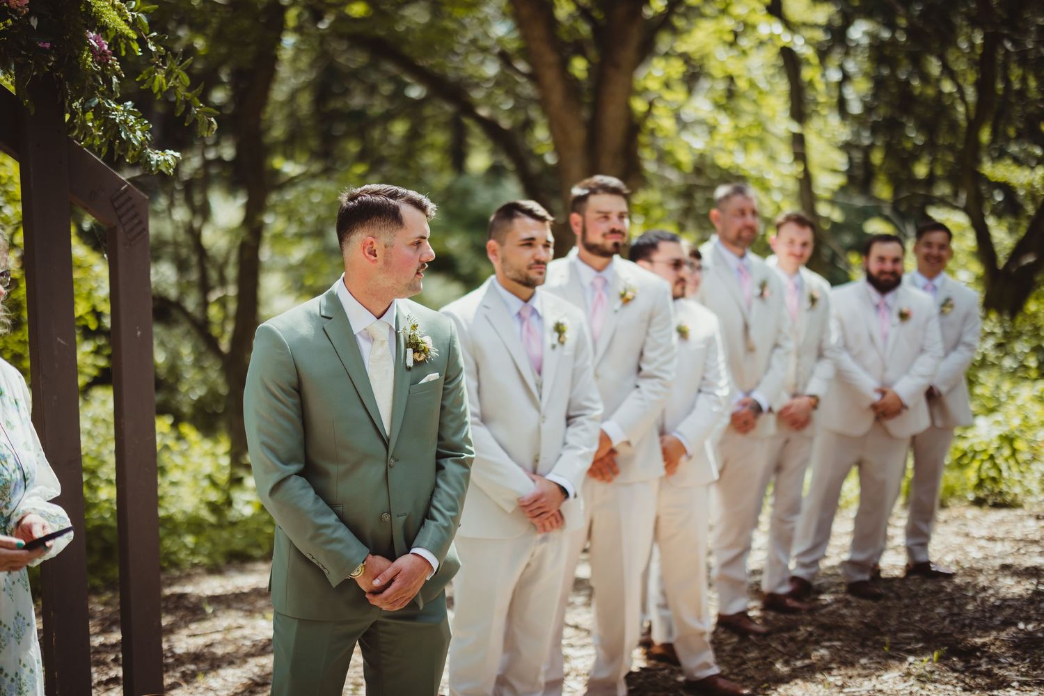 Groom in sage green suit stands with groomsmen in ivory suits lined up outdoors at a woodland wedding ceremony.