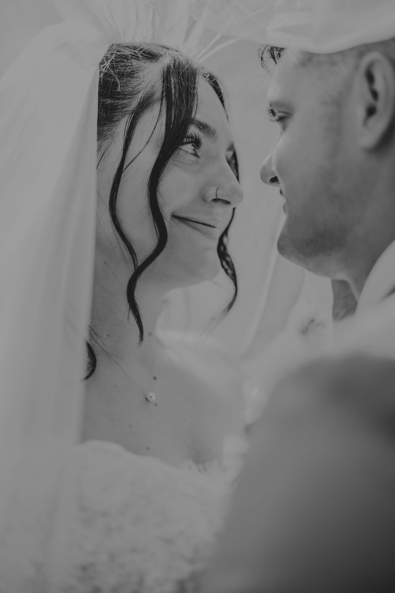 Black and white wedding photo of bride smiling up at groom, veil draped around them in intimate moment.