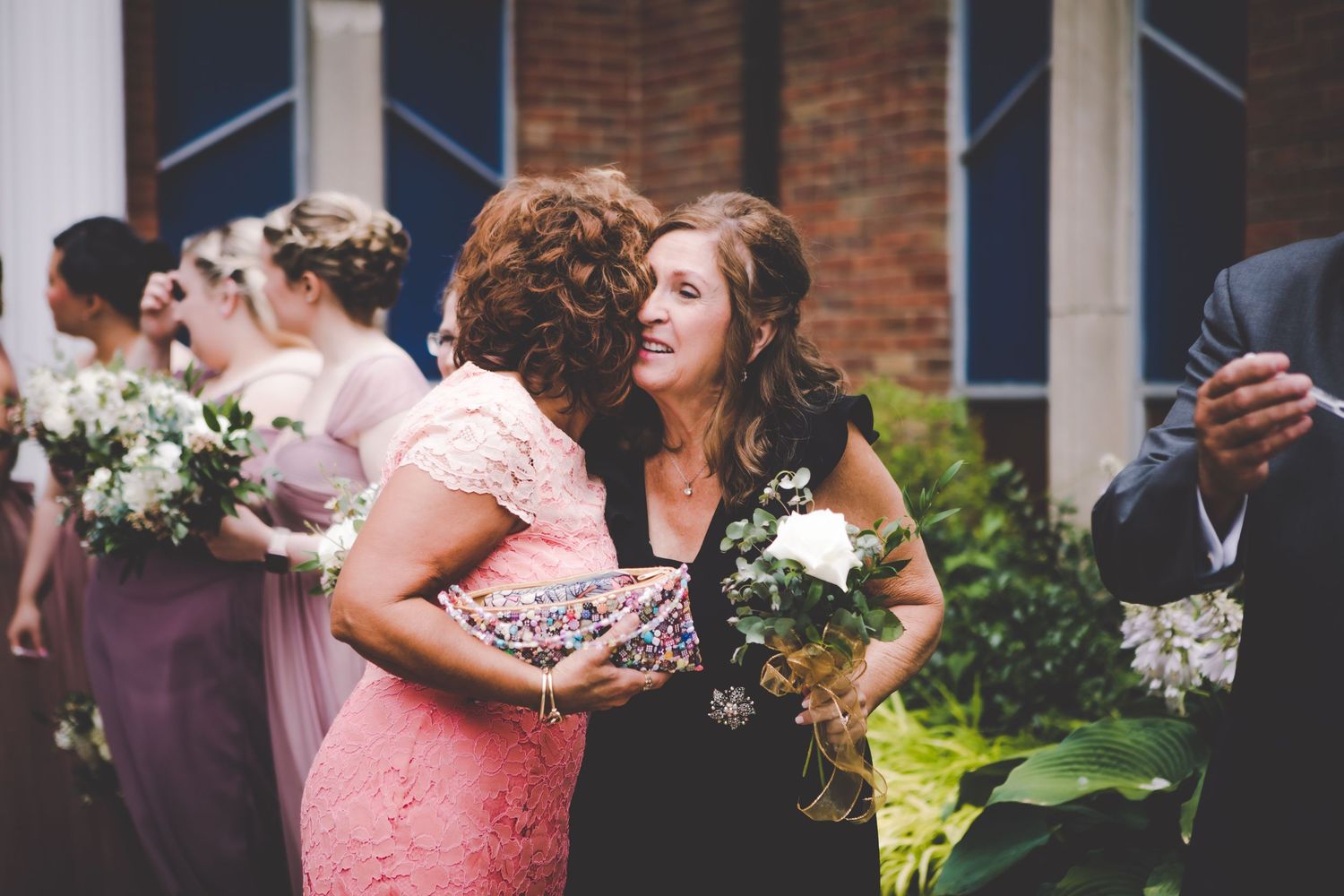 Wedding guests in elegant attire embrace and hold bouquets during an outdoor celebration.