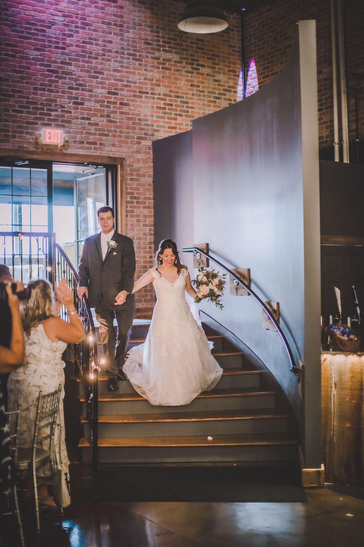Wedding couple descends elegant staircase in industrial venue with exposed brick walls and ambient lighting.