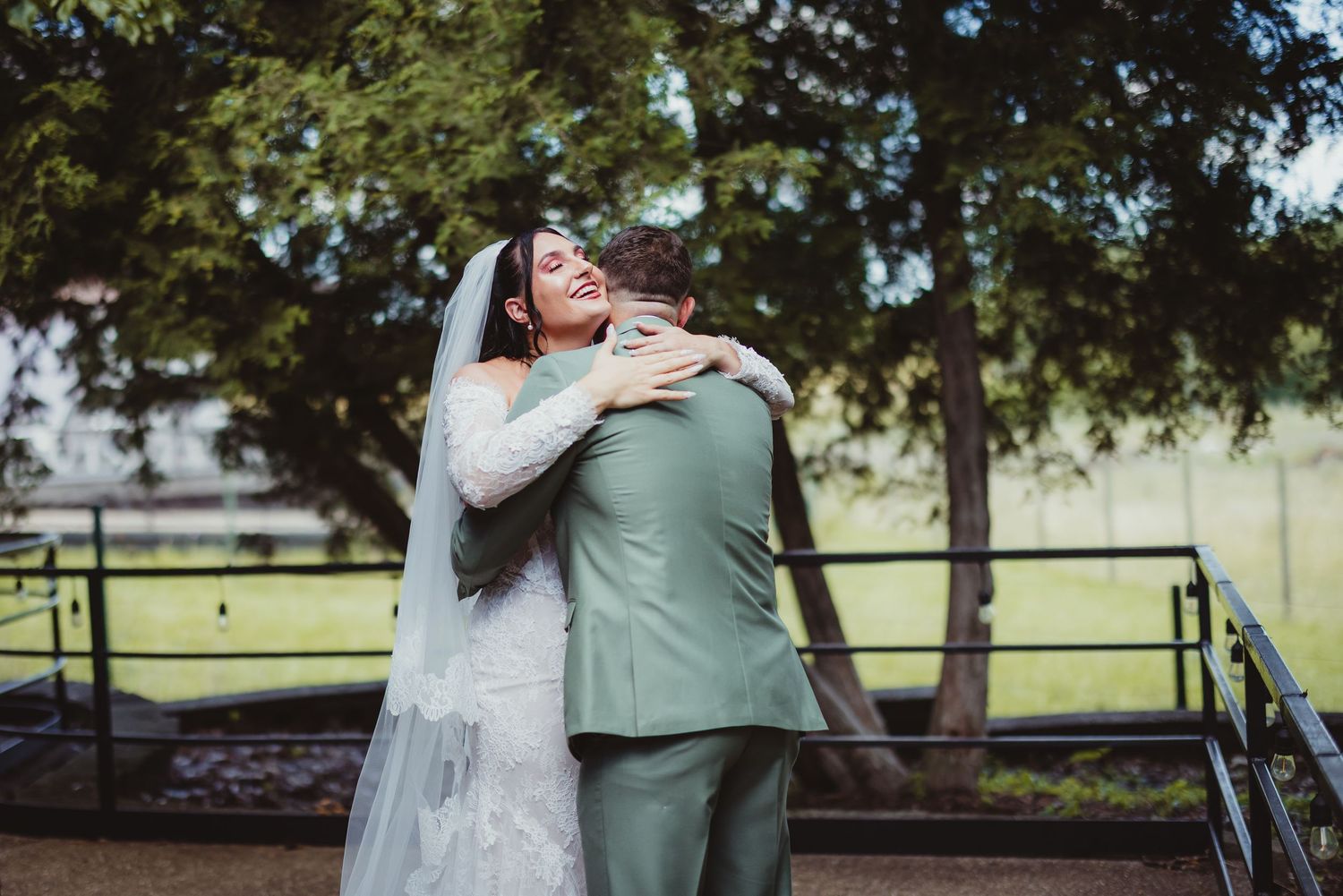 Bride in white gown and veil hugging groom in sage green suit outdoors near trees at wedding celebration.