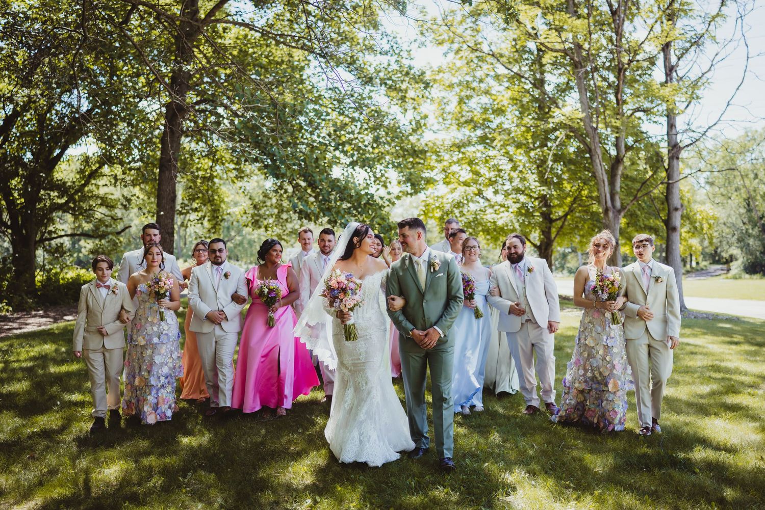 Wedding party in colorful attire poses outdoors on green lawn with trees, bride and groom centered in white.