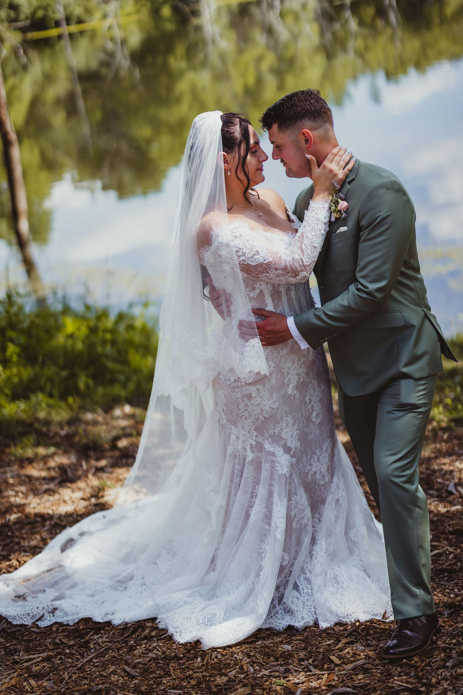 Bride in lace gown and veil with groom in sage suit sharing romantic moment by a lake.