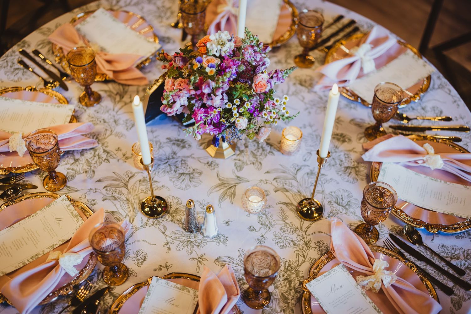 Elegant wedding table setting with gold accents, pink napkins, floral centerpiece, white candles, and toile tablecloth.