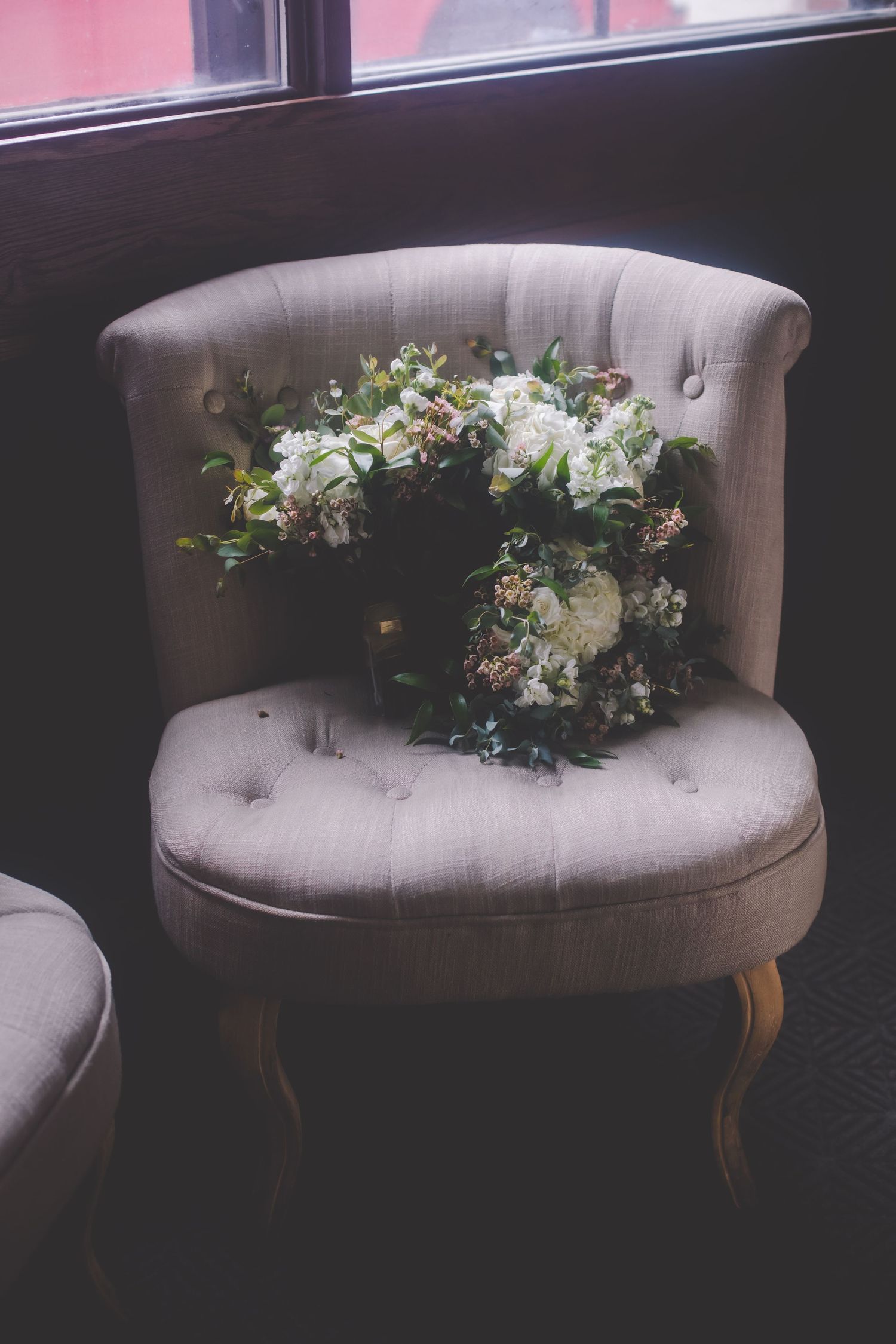 Vintage gray tufted chair adorned with white floral wreath in window light.