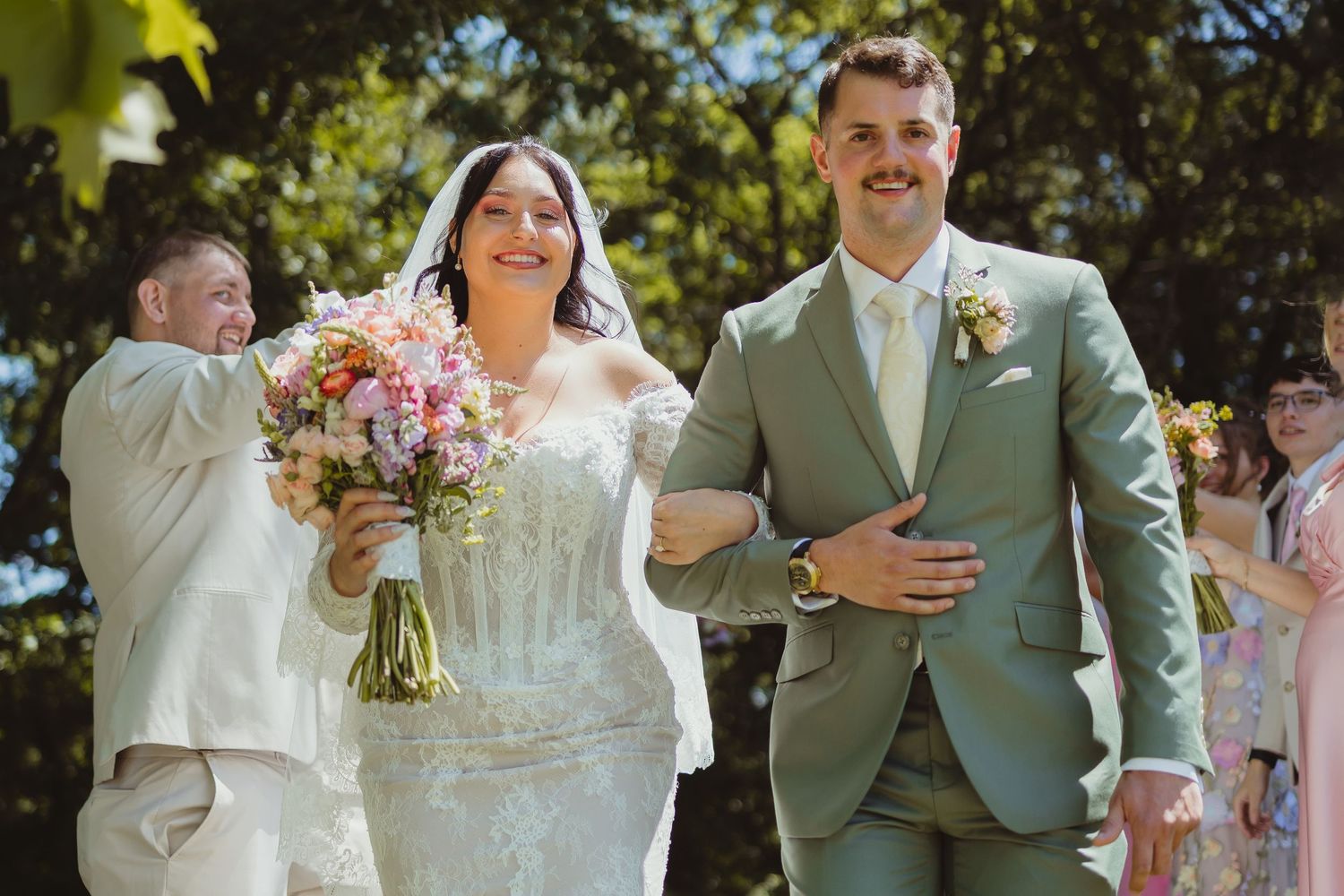 Smiling bride holding colorful bouquet walks arm-in-arm with groom in sage suit at outdoor wedding ceremony.