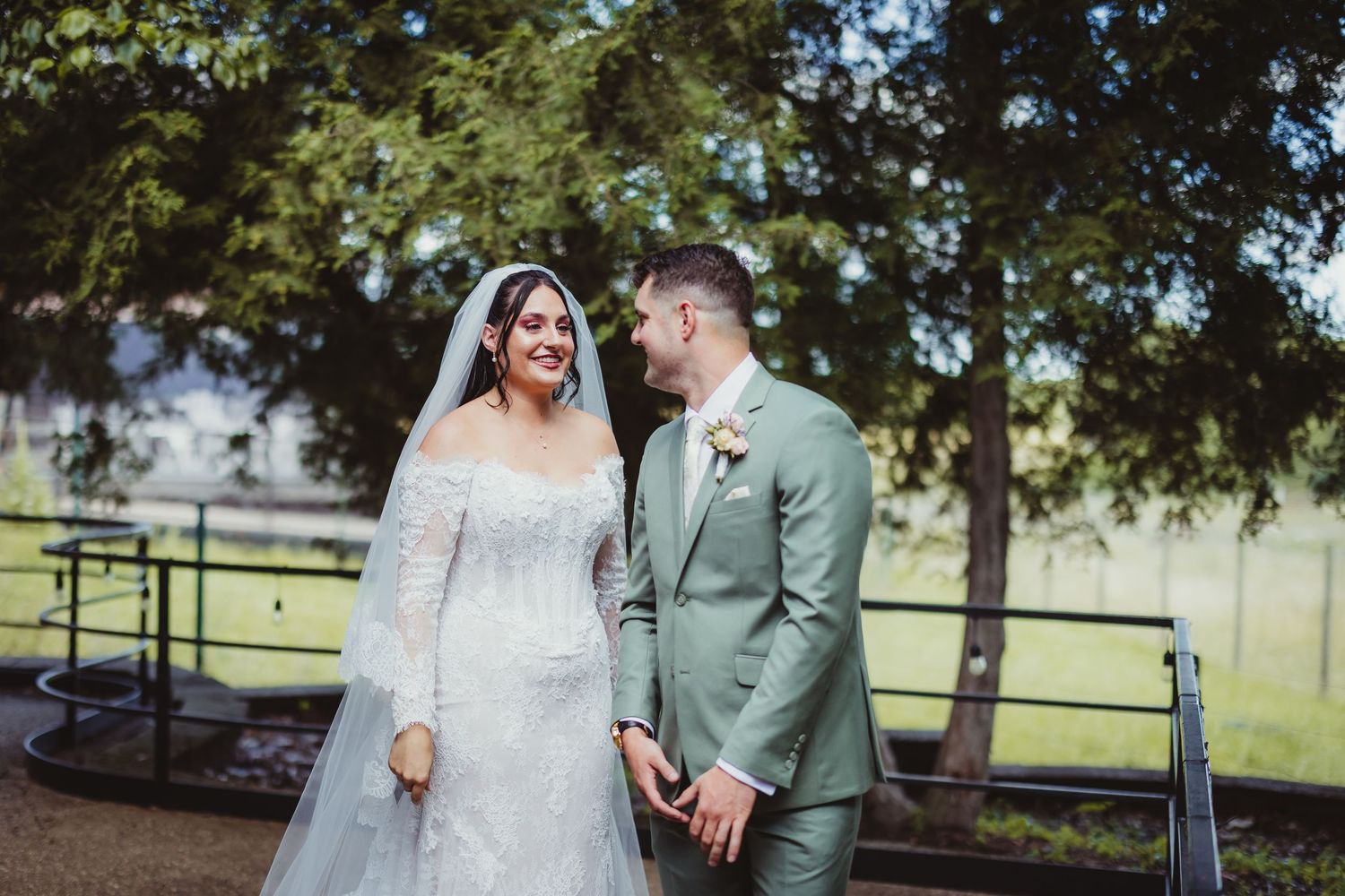 Bride in lace gown and groom in sage suit hold hands smiling at each other at outdoor wedding.