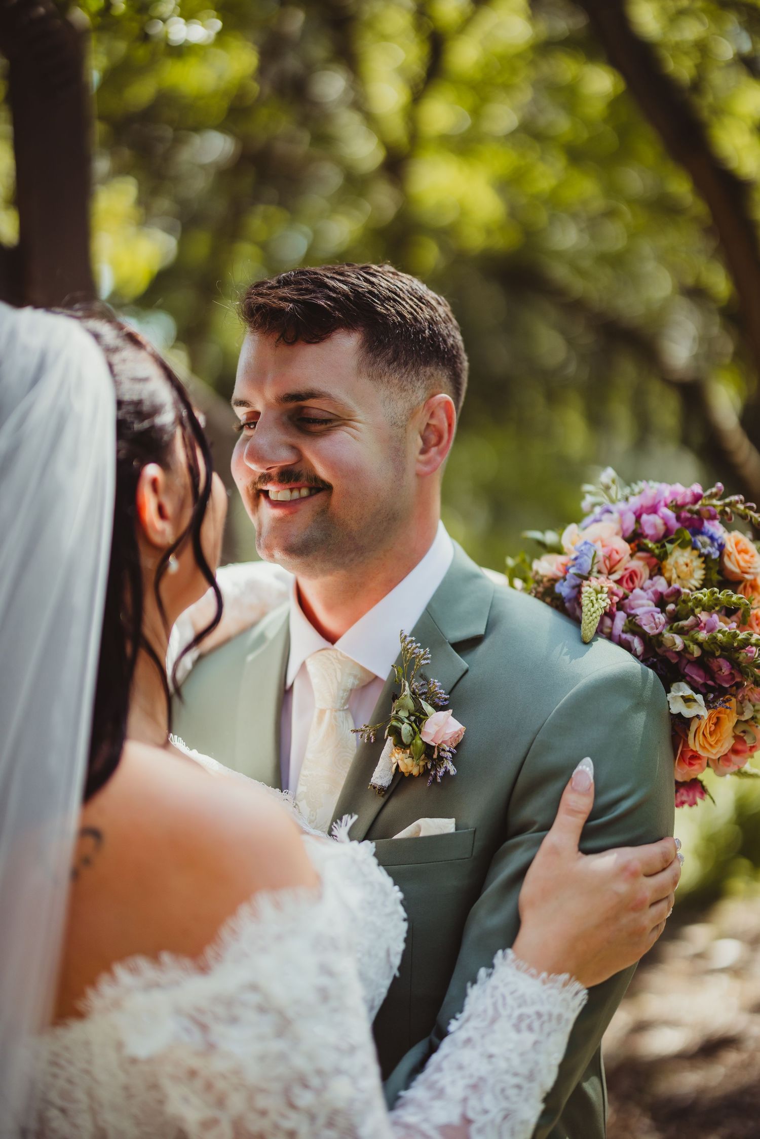 Groom in sage green suit smiling at bride holding colorful bouquet during outdoor wedding ceremony.