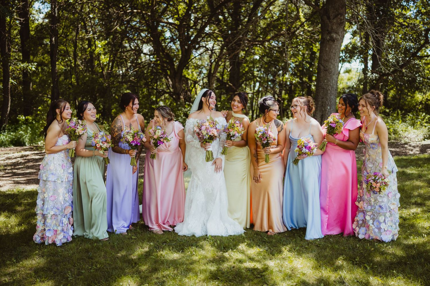 Bride in white gown poses with colorful rainbow bridesmaid dresses in lush green outdoor wedding setting.