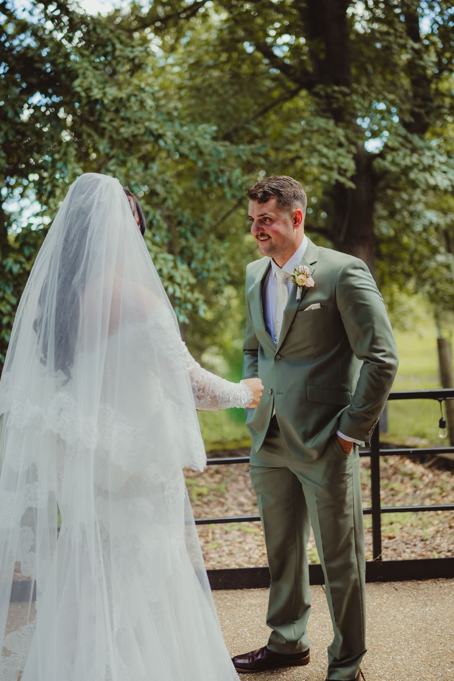 Groom in sage green suit smiles at bride in white lace gown and veil during outdoor first look ceremony.