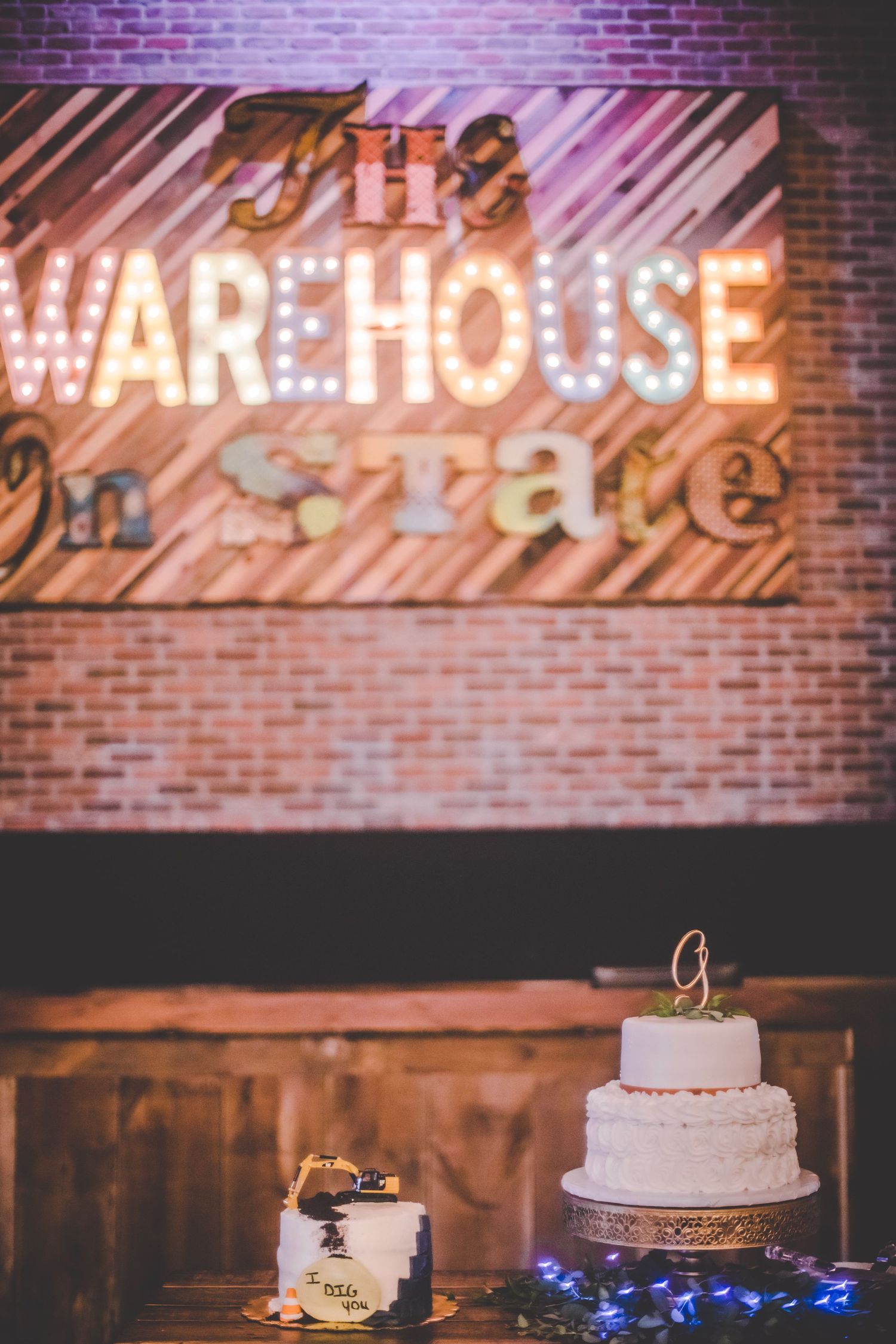 A two-tiered wedding cake displayed beneath an illuminated 'The Warehouse' sign on a brick wall.