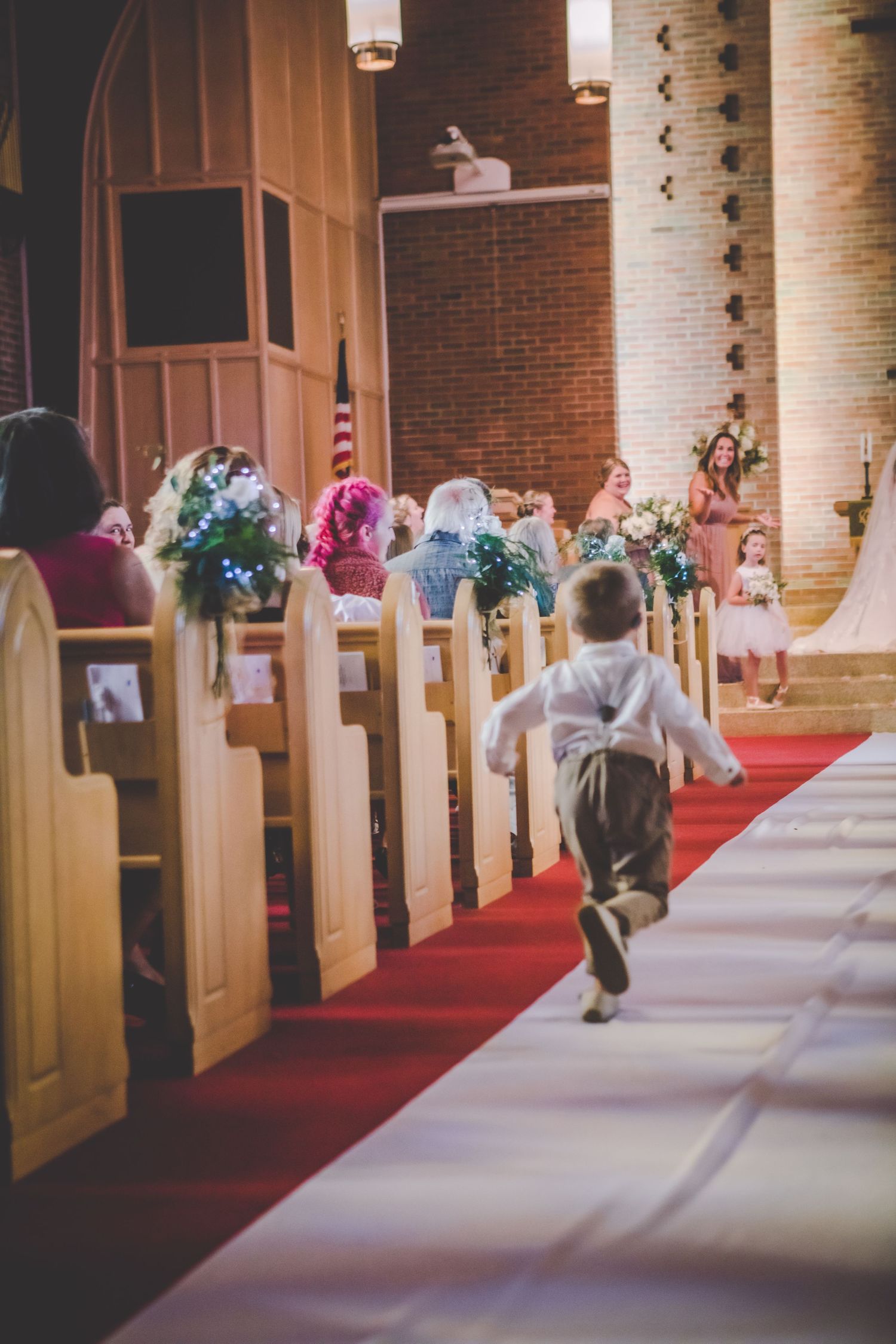 Child running down red-carpeted church aisle during wedding ceremony with guests seated in pews.