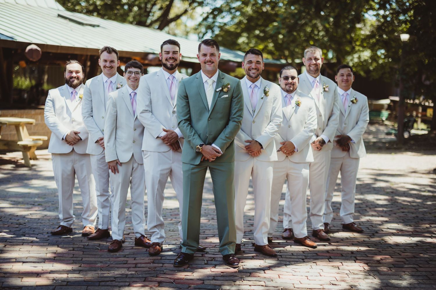 Groom in sage green suit poses with groomsmen in white suits outdoors at a rustic wedding venue.