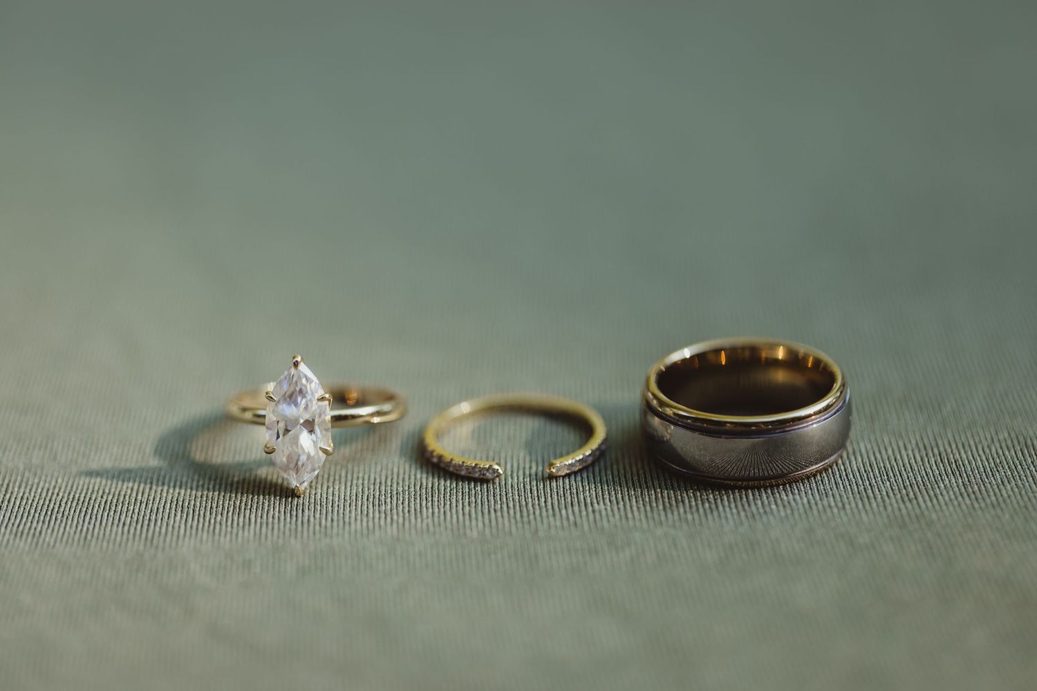 Three wedding rings on green fabric: diamond engagement ring, gold band, and two-tone titanium band.