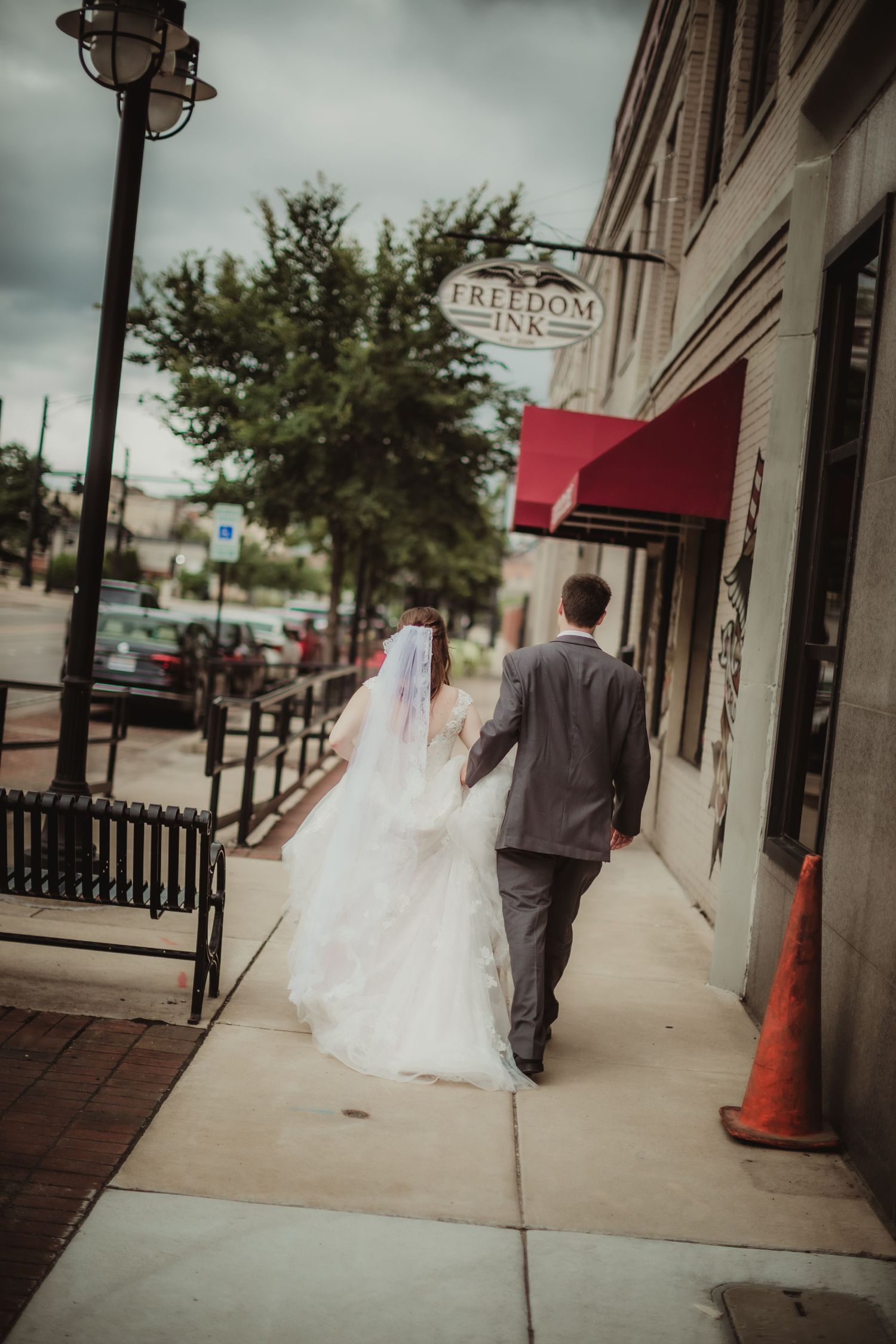 Bride and groom walk down urban sidewalk past storefront with red awning.