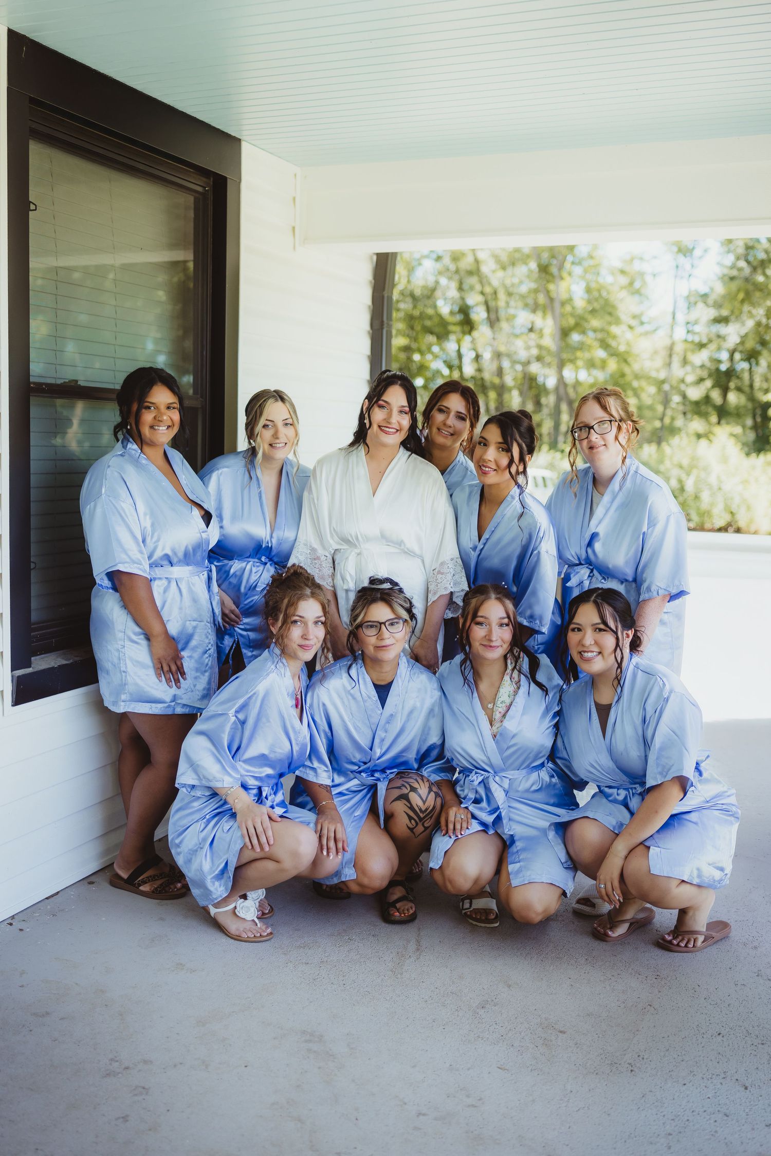 Bride in white robe poses with bridesmaids in matching blue robes on a porch during wedding day preparations.