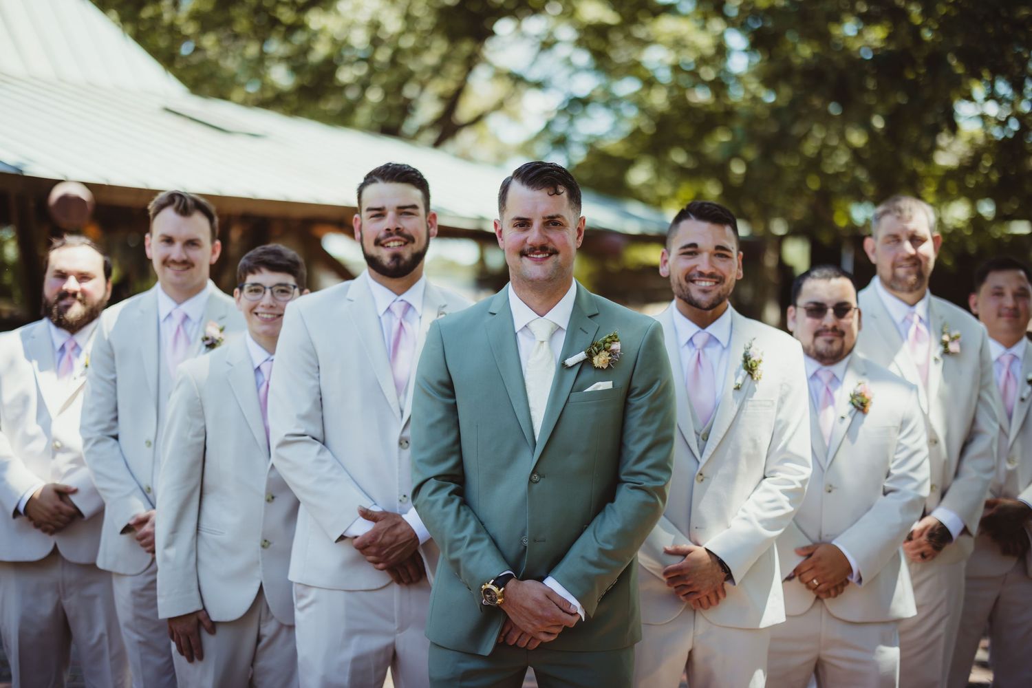 Groom in mint green suit stands with groomsmen in light gray suits and lavender ties at outdoor wedding.