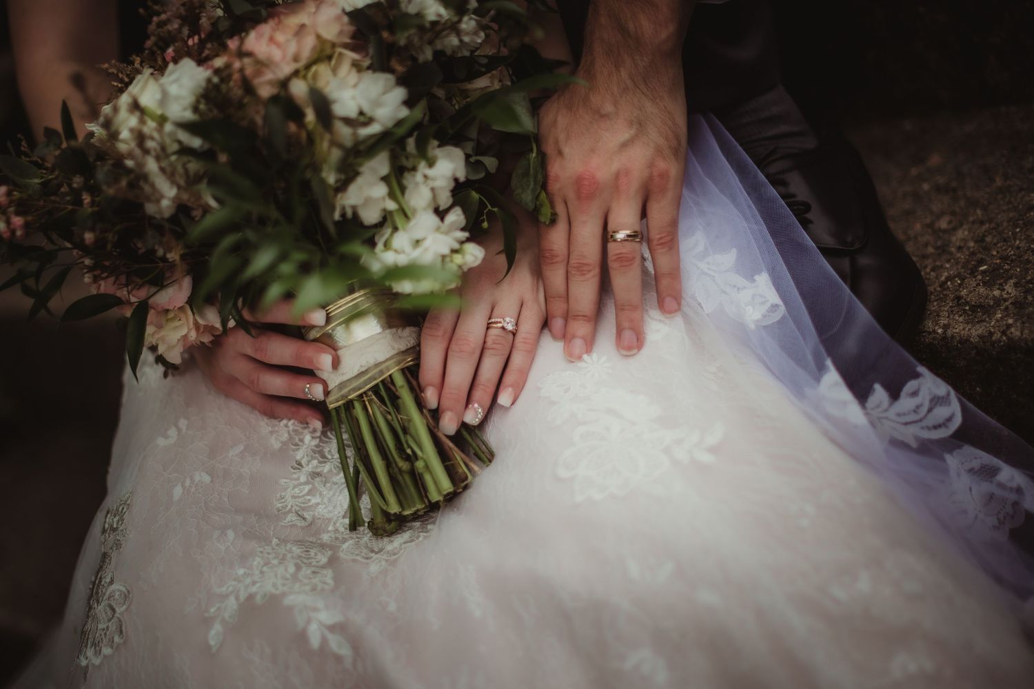 Close-up of wedding rings and bouquet against white lace dress fabric.