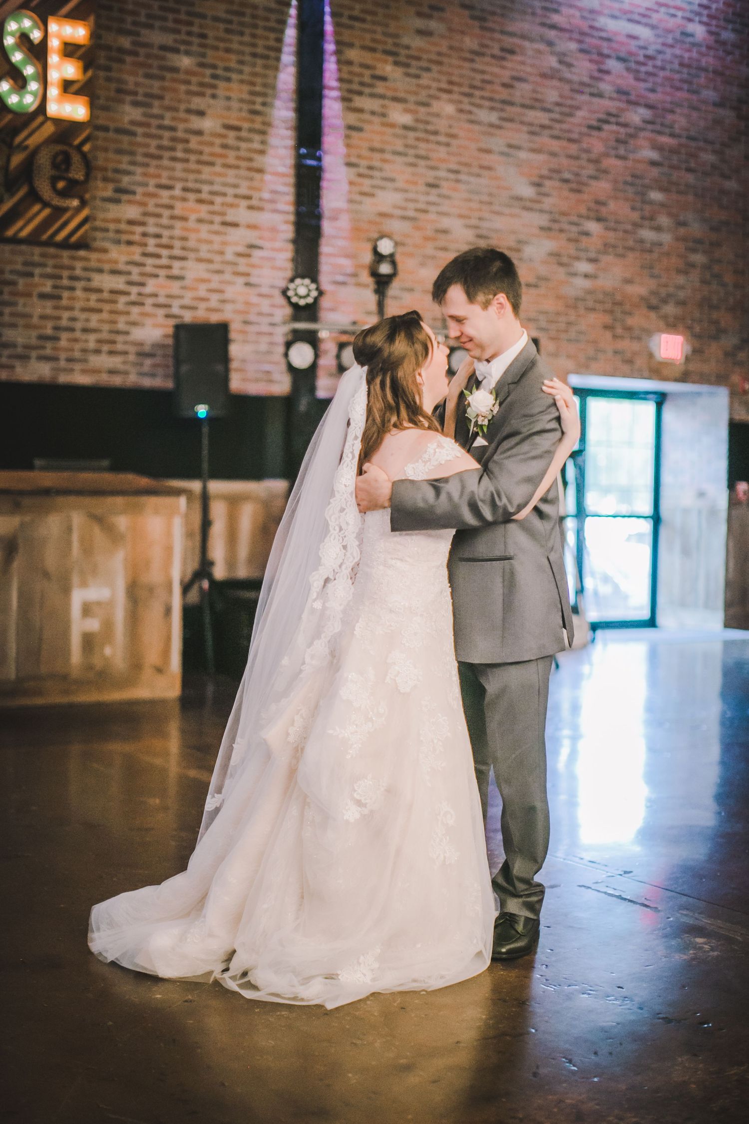 A couple in wedding attire shares a romantic dance in a rustic brick-walled venue.