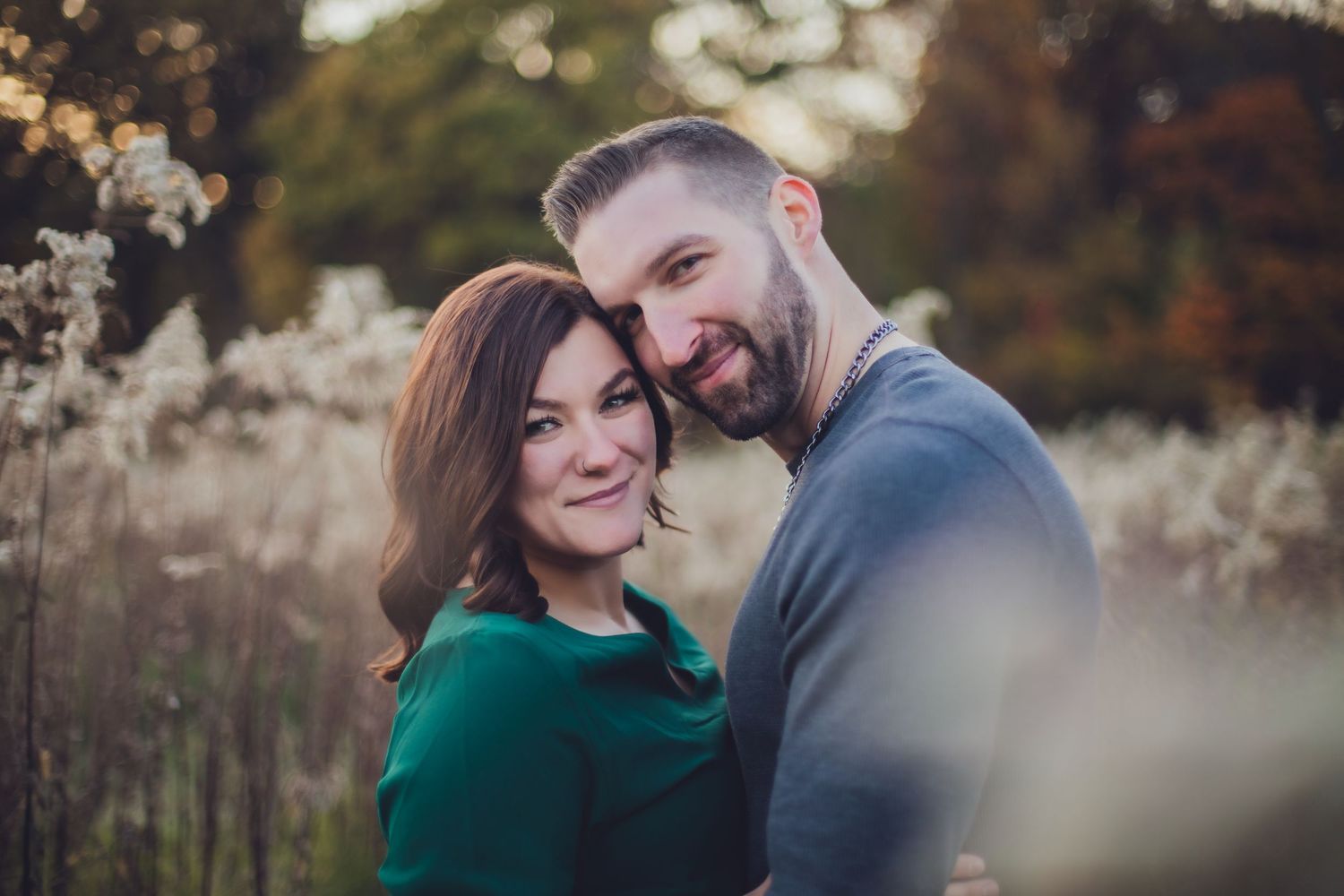 A couple embraces in soft focus among autumn grasses wearing green and blue casual attire at sunset.