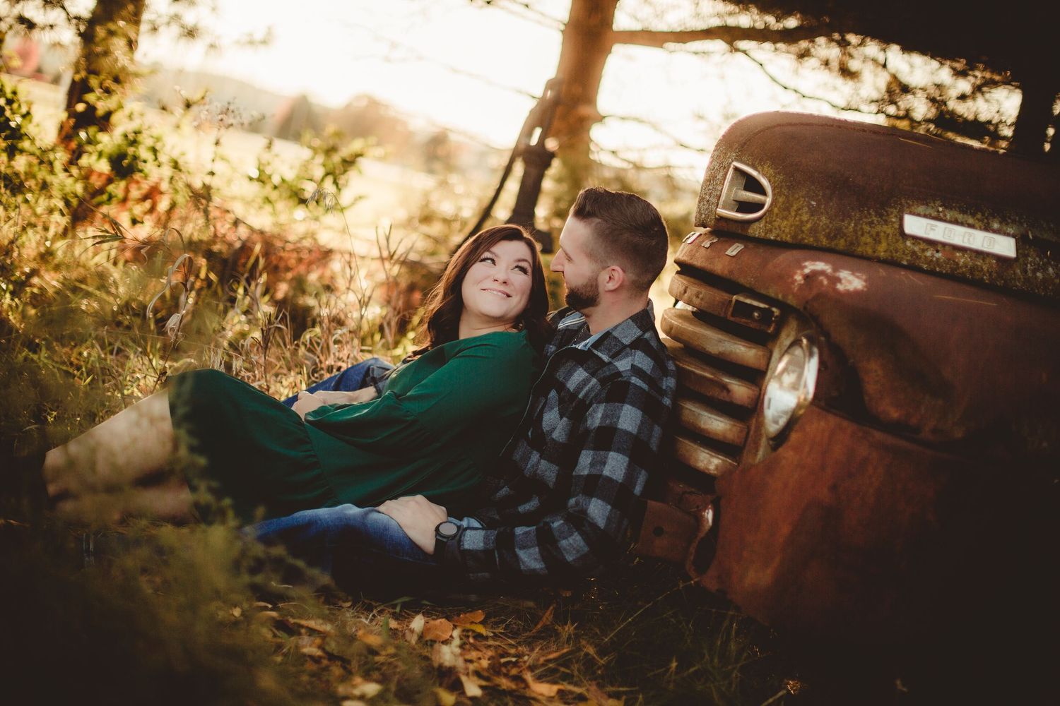 Couple relaxes against an old pickup truck during sunset in autumn setting.