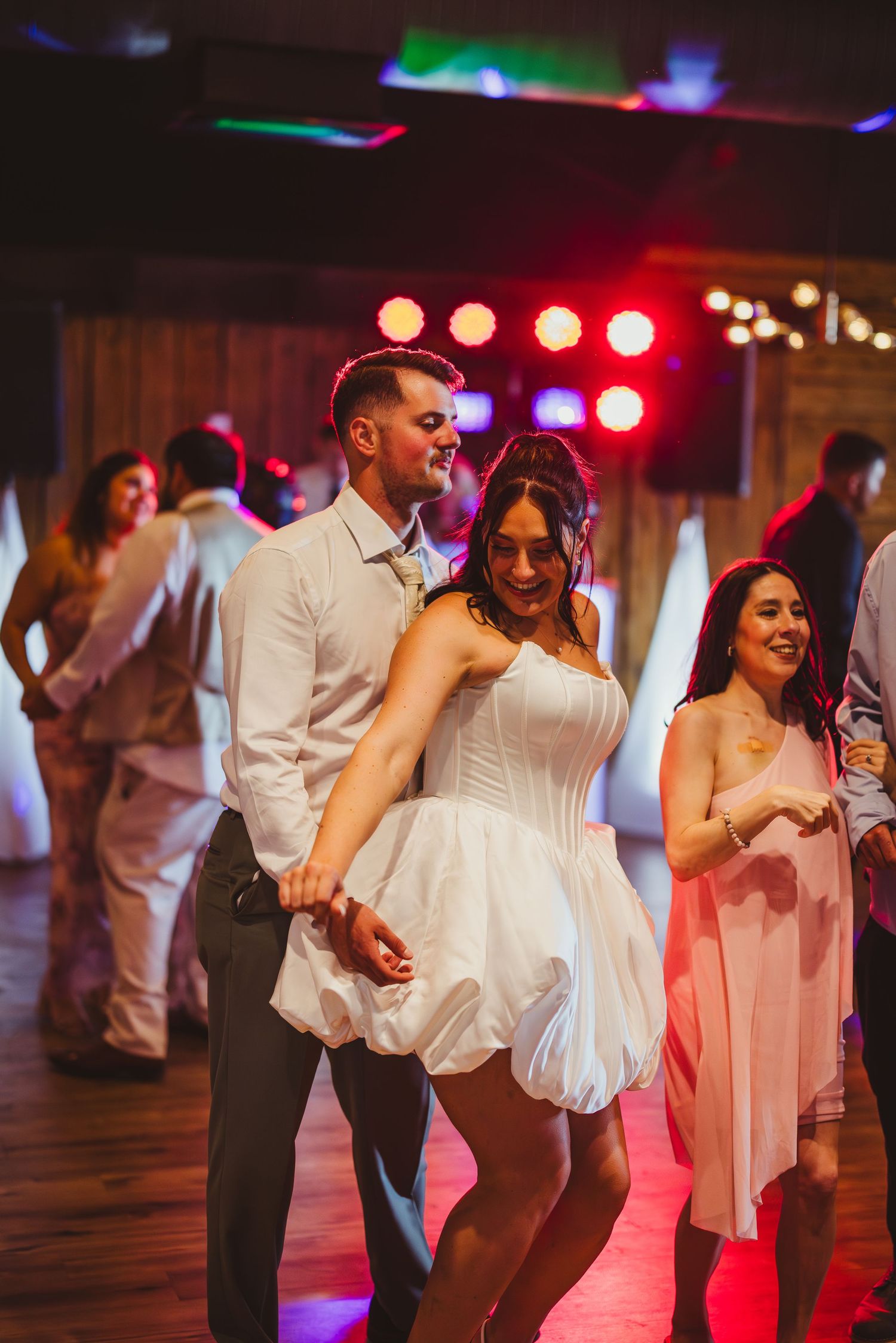 Bride in white bubble dress dancing with groom at wedding reception under colorful stage lights.