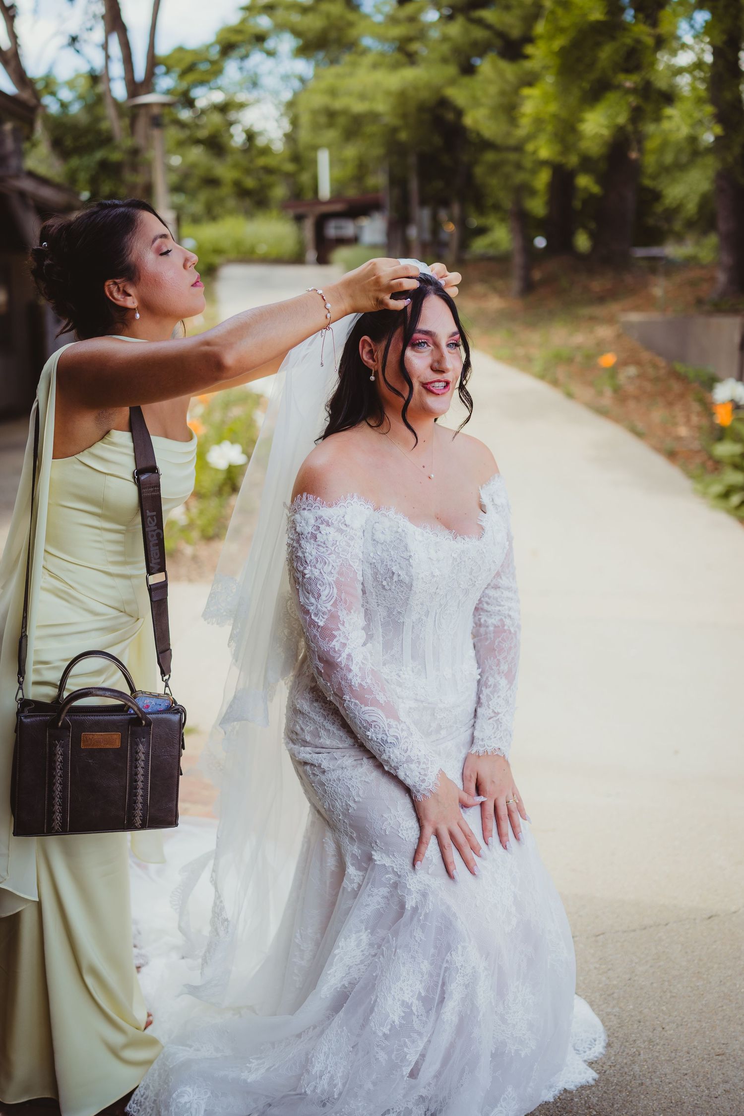 Bridesmaid in sage green dress adjusts veil on smiling bride in lace off-shoulder wedding gown outdoors.