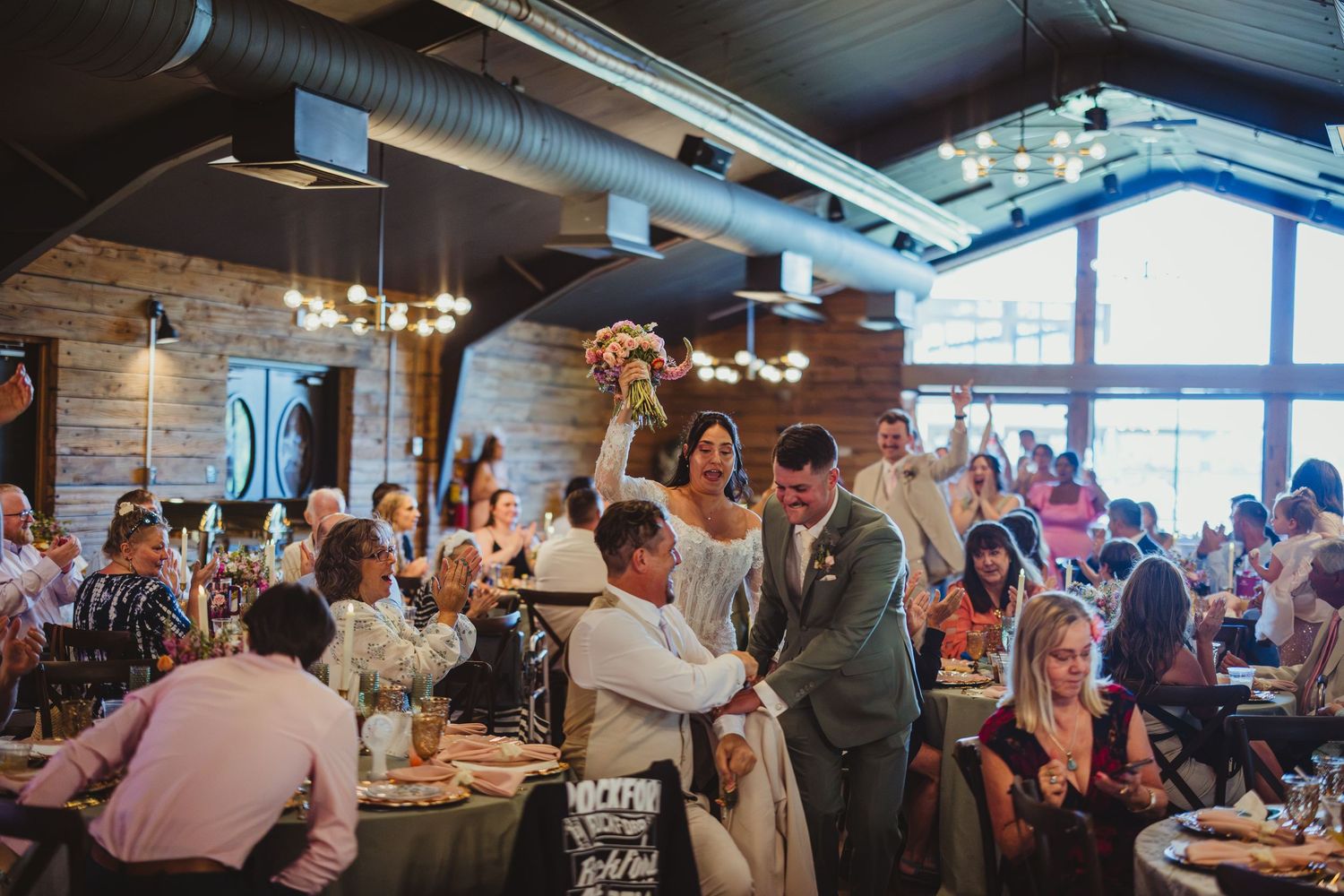 Bride and groom walk triumphantly through wedding reception hall filled with guests at decorated tables.