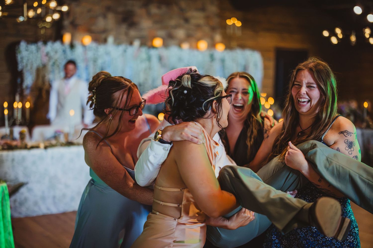 Bridesmaids joyfully lifting the bride during a lively wedding reception celebration in a rustic venue.