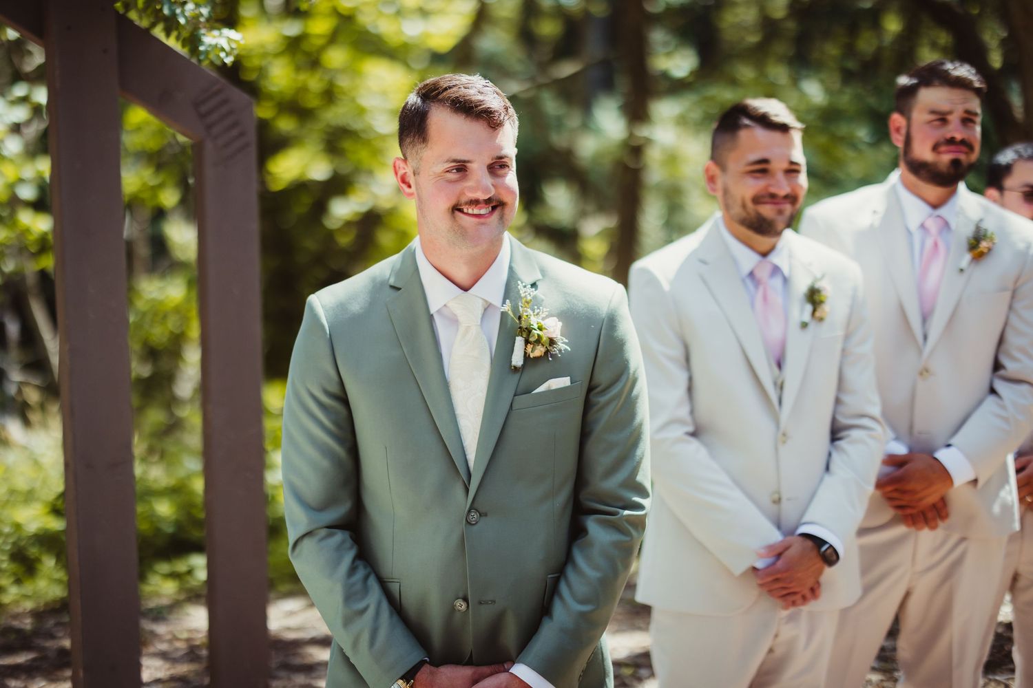 Smiling groom in sage green suit stands at altar with groomsmen in beige suits during outdoor wedding ceremony.